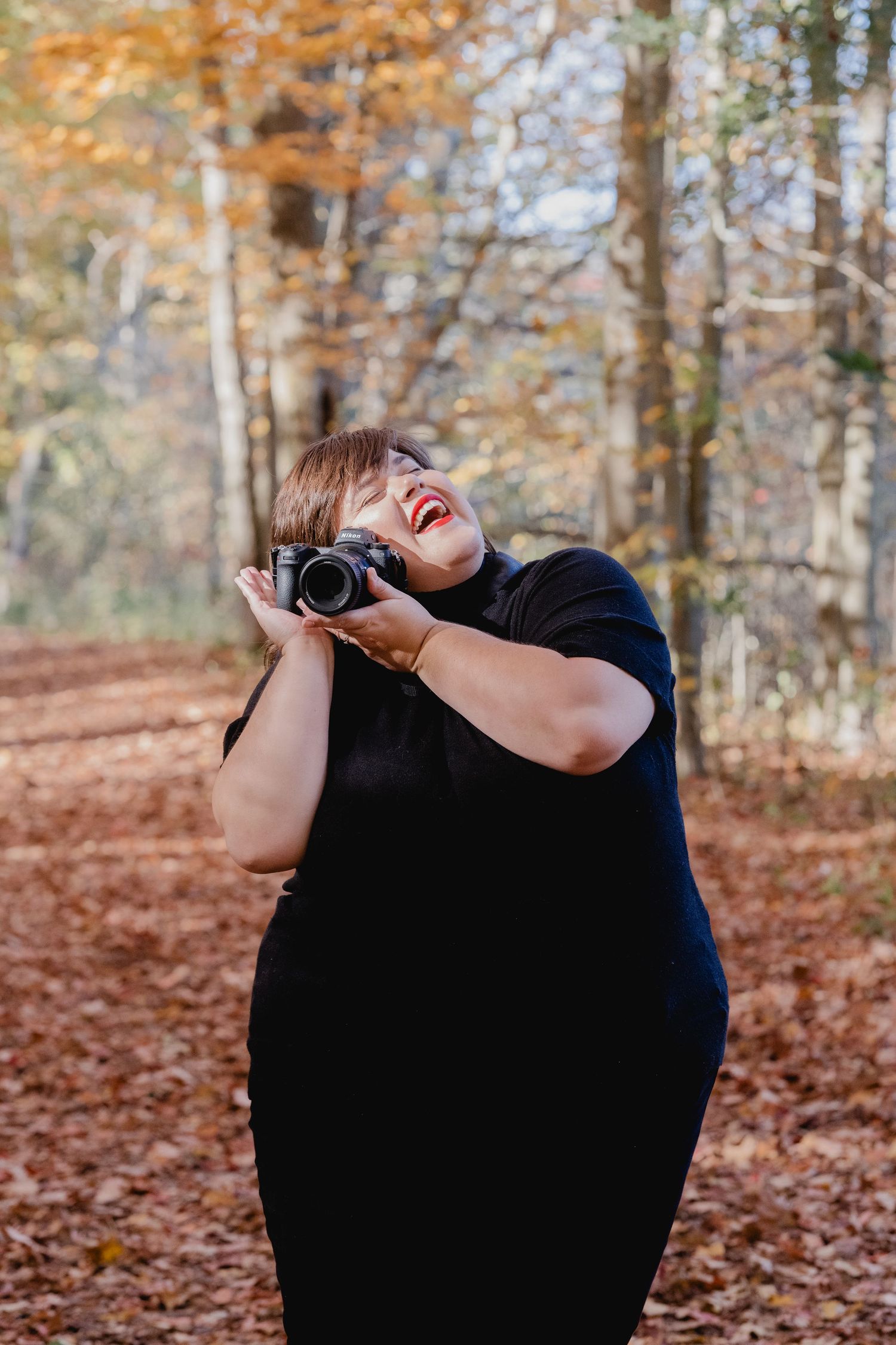 Someone wearing a black dress laughs joyfully while holding a camera in an autumn forest with fallen leaves.