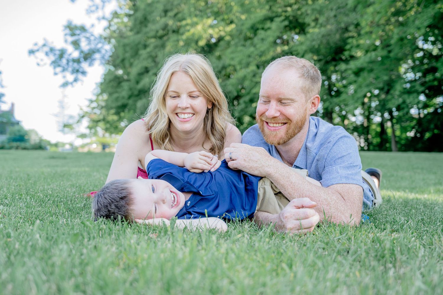 Family of three smiling and laughing together on green grass outdoors, parents lying down with young child between them.