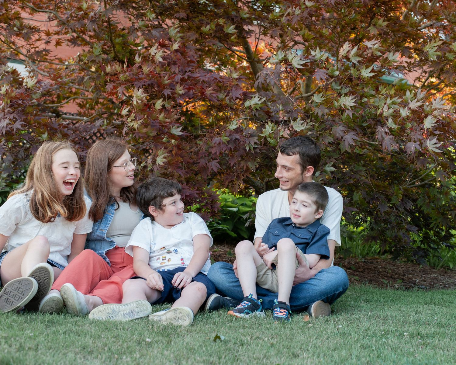 A panoramic view of children relaxing on green grass under autumn foliage at a park.