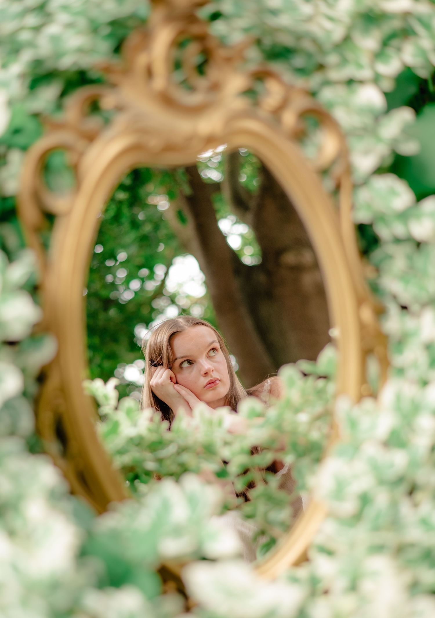 A dreamy spring portrait through an ornate wooden archway with soft bokeh and blossoming white flowers in the background.