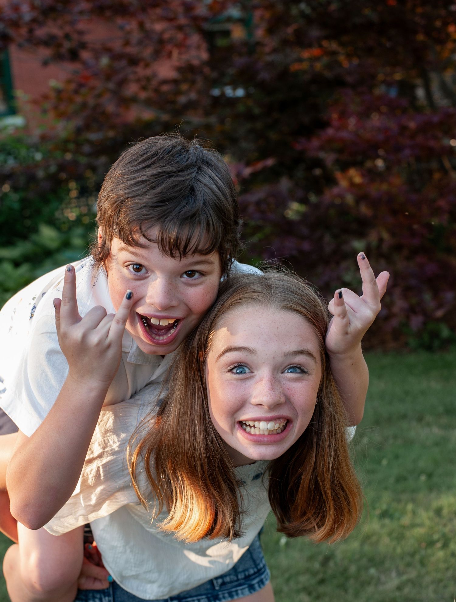Two children making silly expressions and playful gestures together outdoors with autumn foliage in background.