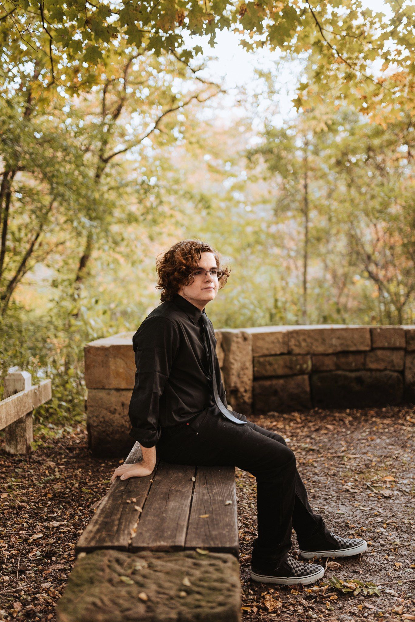 A person in black clothing sits thoughtfully on a rustic wooden bench surrounded by autumn leaves and stone walls.