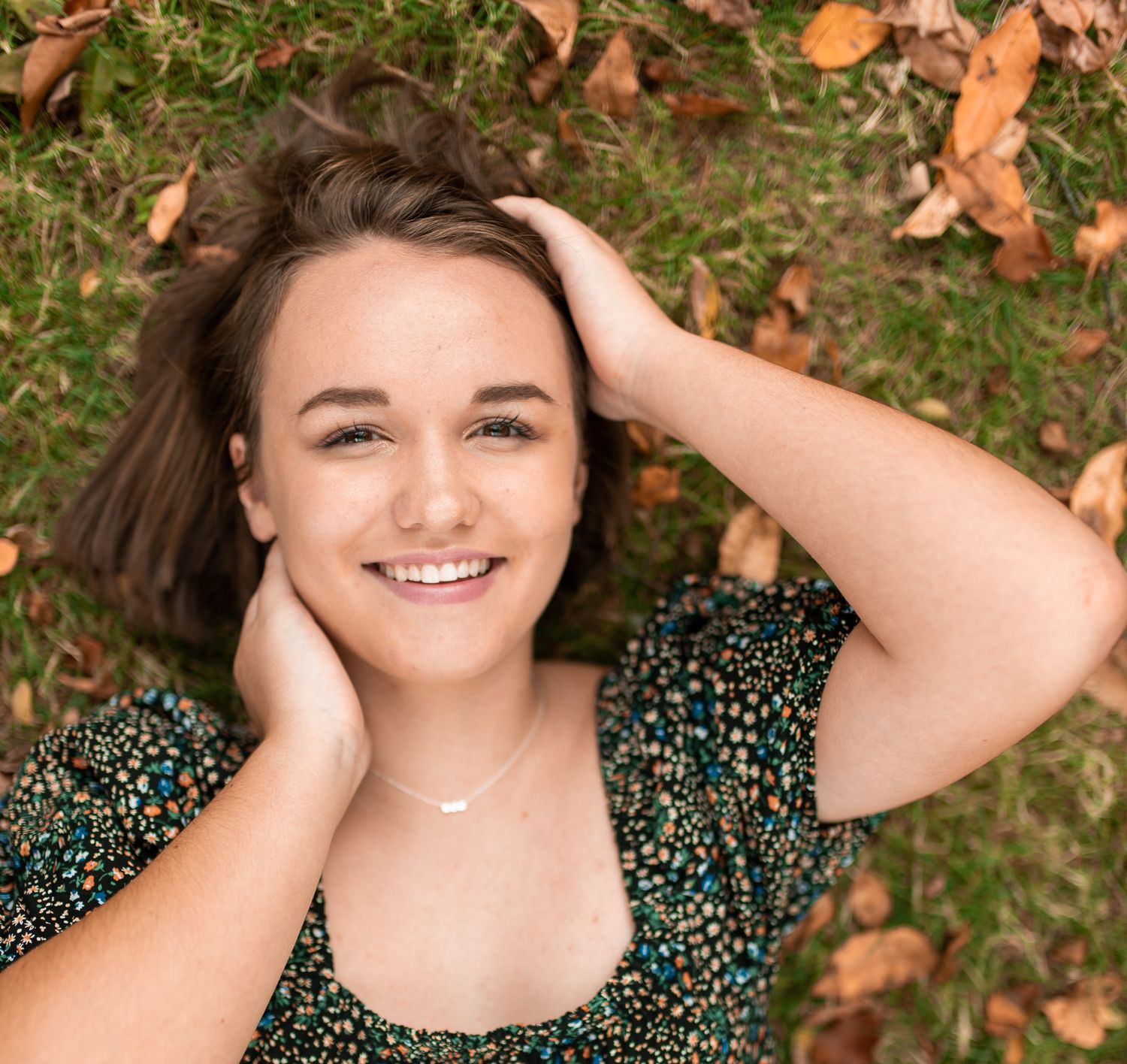 Person in polka dot dress smiling while lying on autumn leaves in the grass during an outdoor photo session.