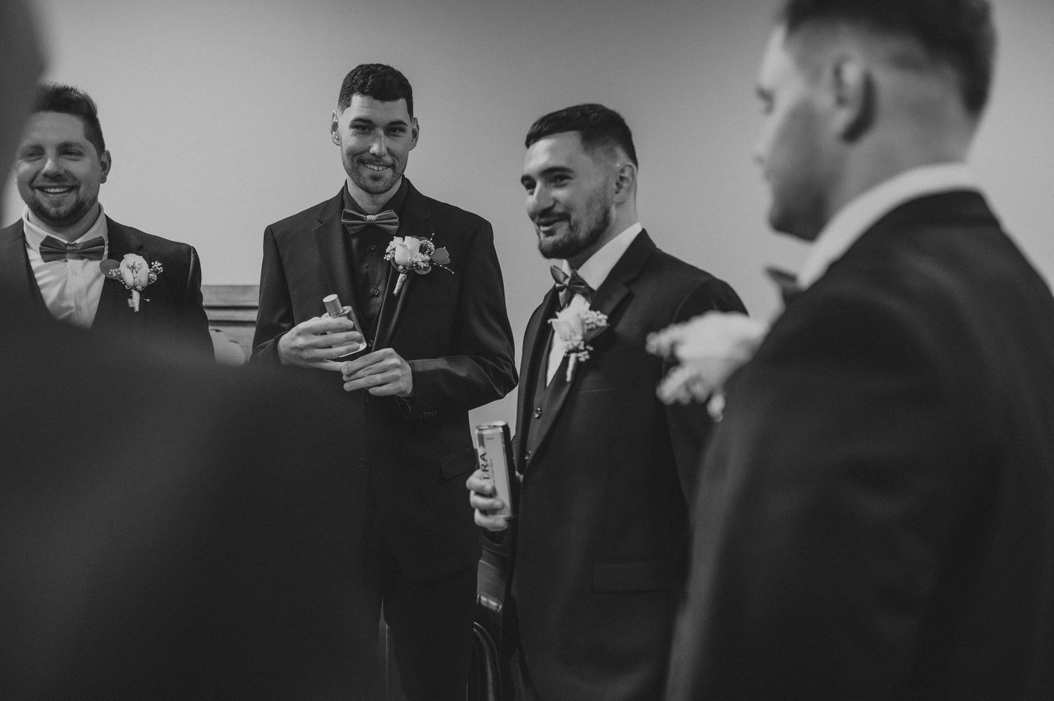 Groomsmen in black suits share drinks and laughs during a wedding celebration.