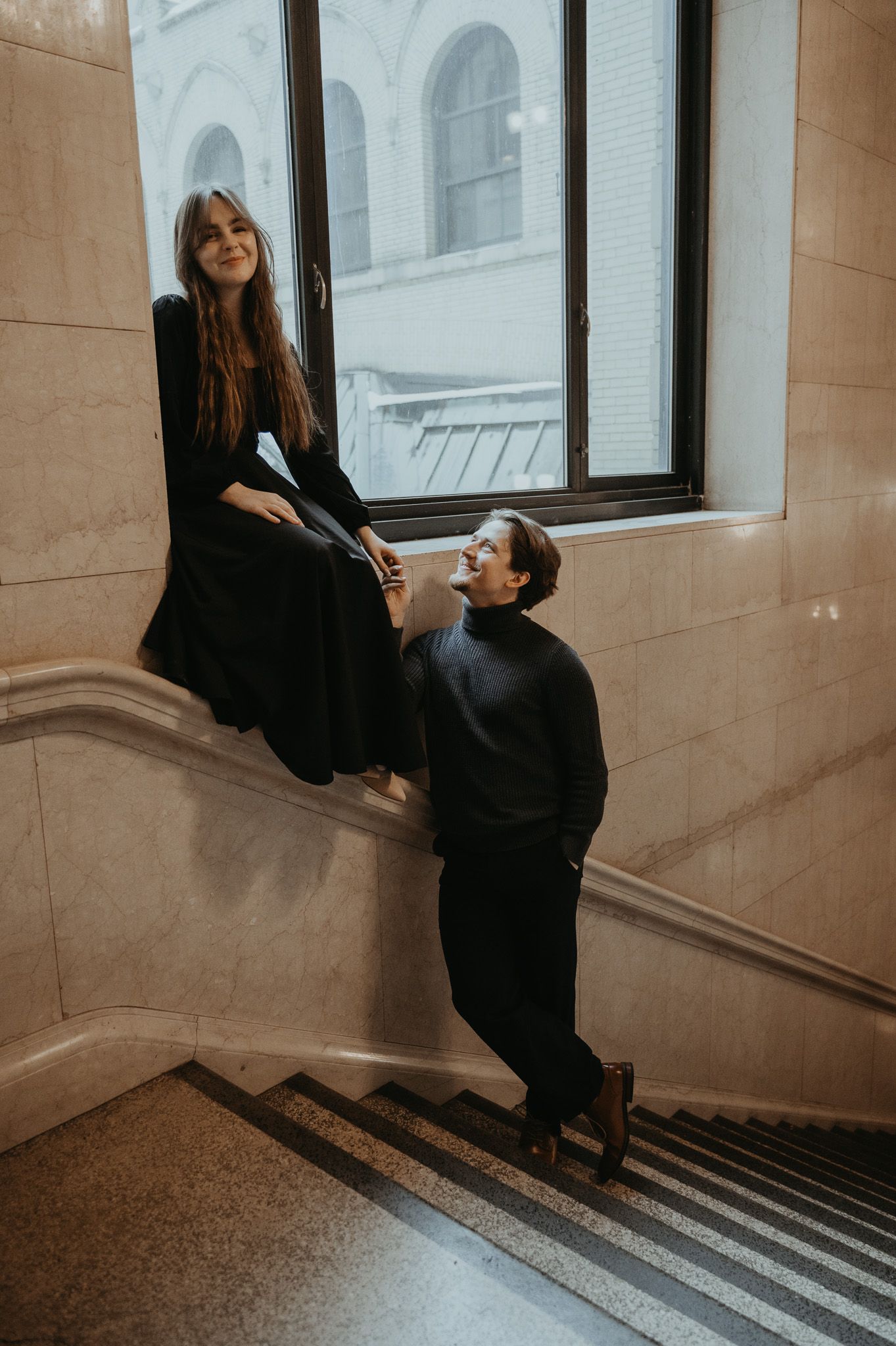 Woman in black outfit sitting on marble staircase in elegant building interior with natural lighting through windows.
