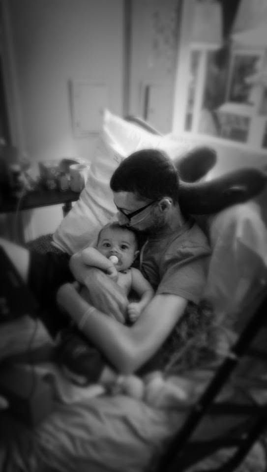 Black and white photo of a father tenderly holding his newborn baby with a pacifier while sitting in a hospital chair.