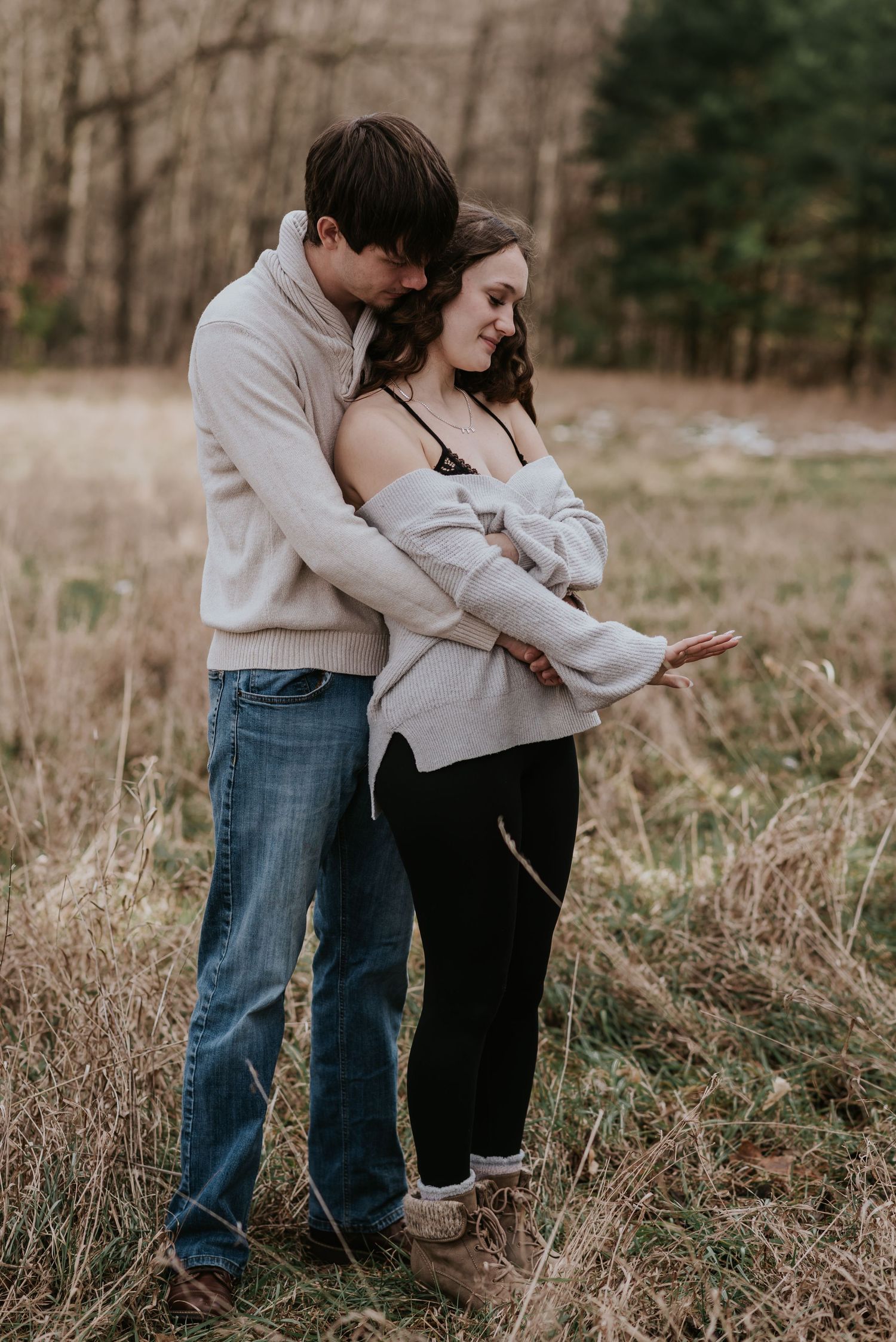 Couple in matching light sweaters embrace while standing in a rustic field.