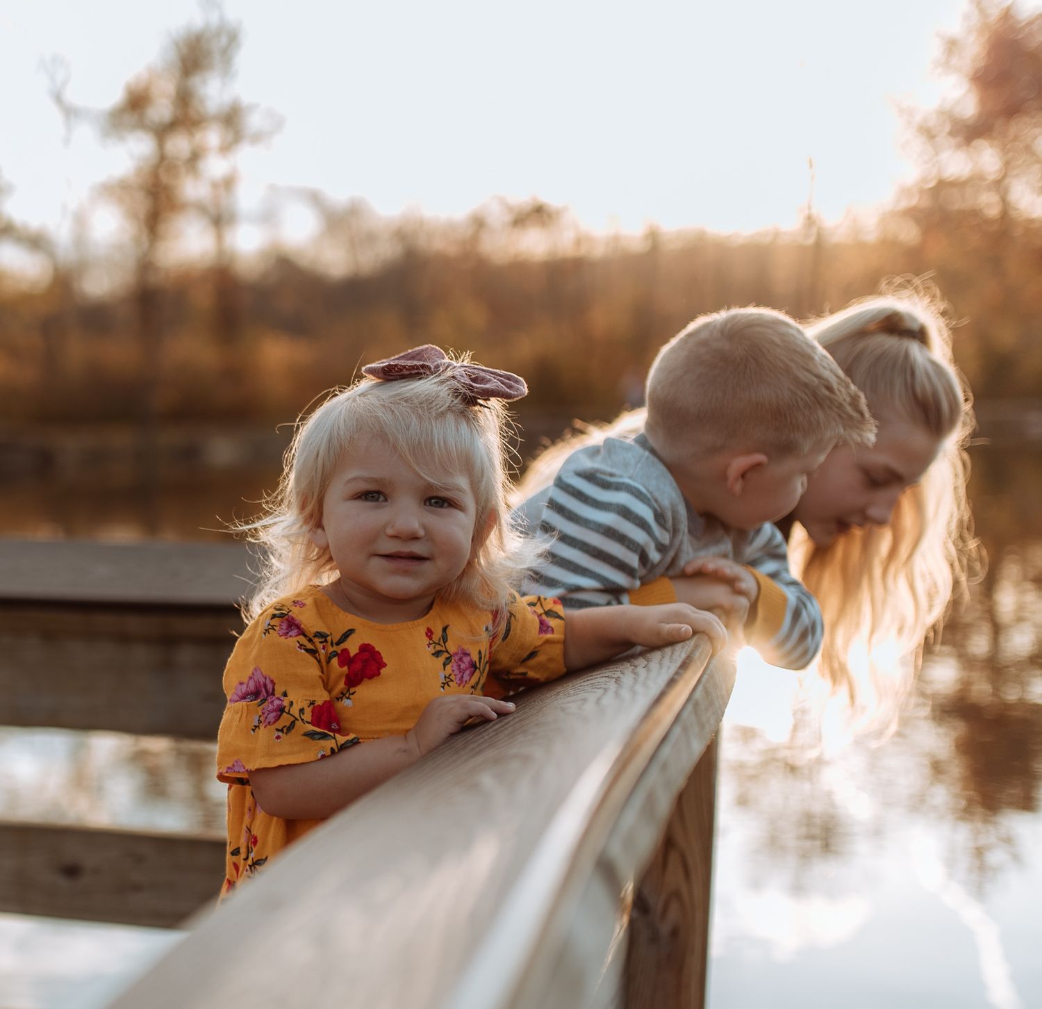 Children lean against a wooden railing overlooking a serene lake at sunset.