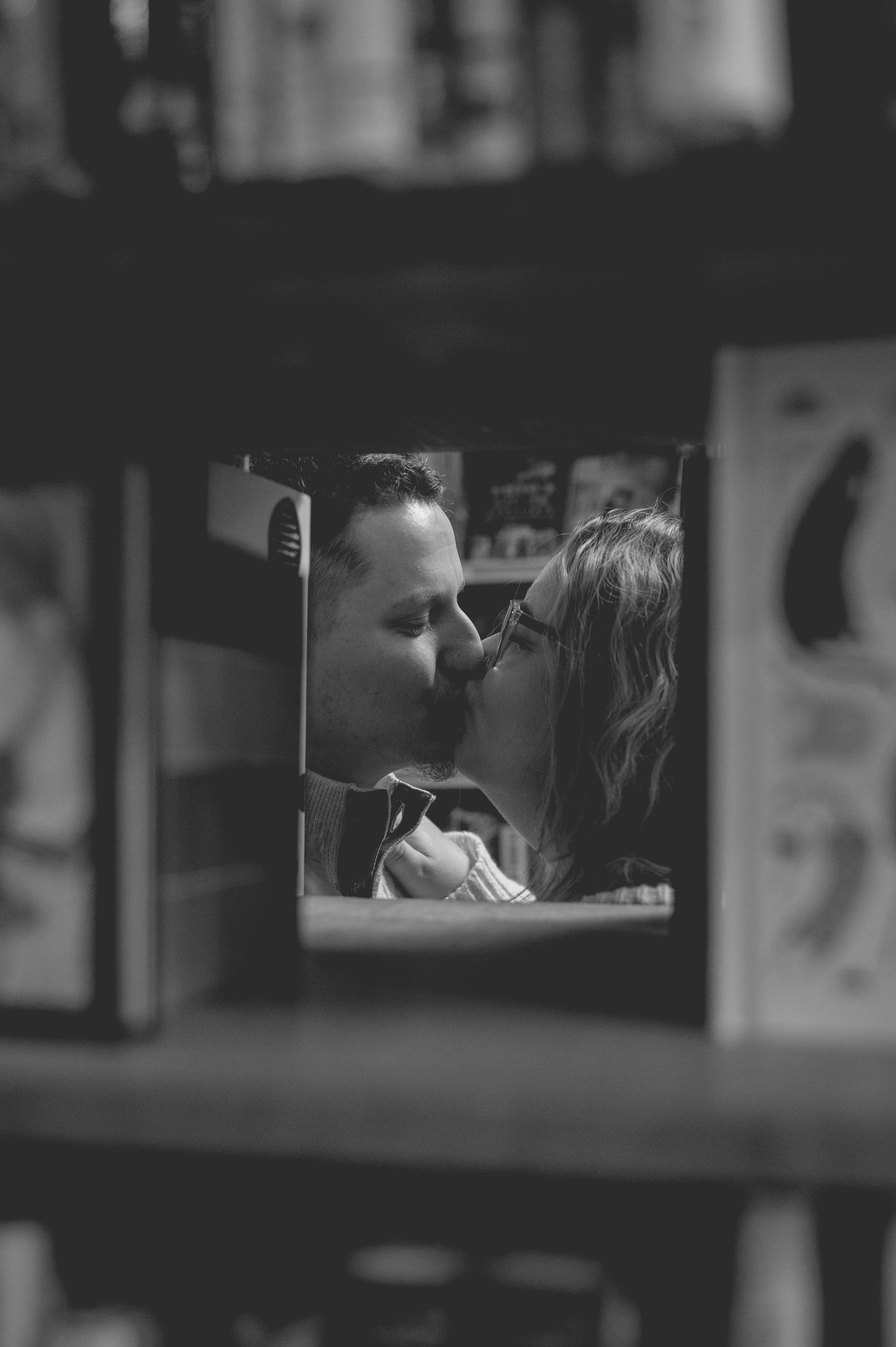Black and white photo of a couple sharing an intimate kiss in a dimly lit room with bookshelves in the background.