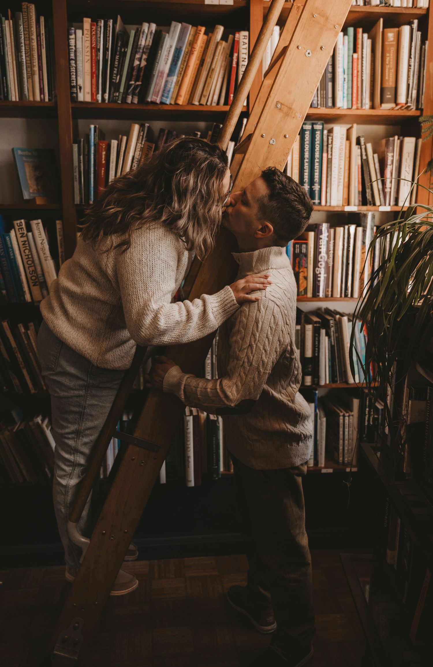 Two people embrace in a romantic moment against a backdrop of bookshelves in a cozy library setting.