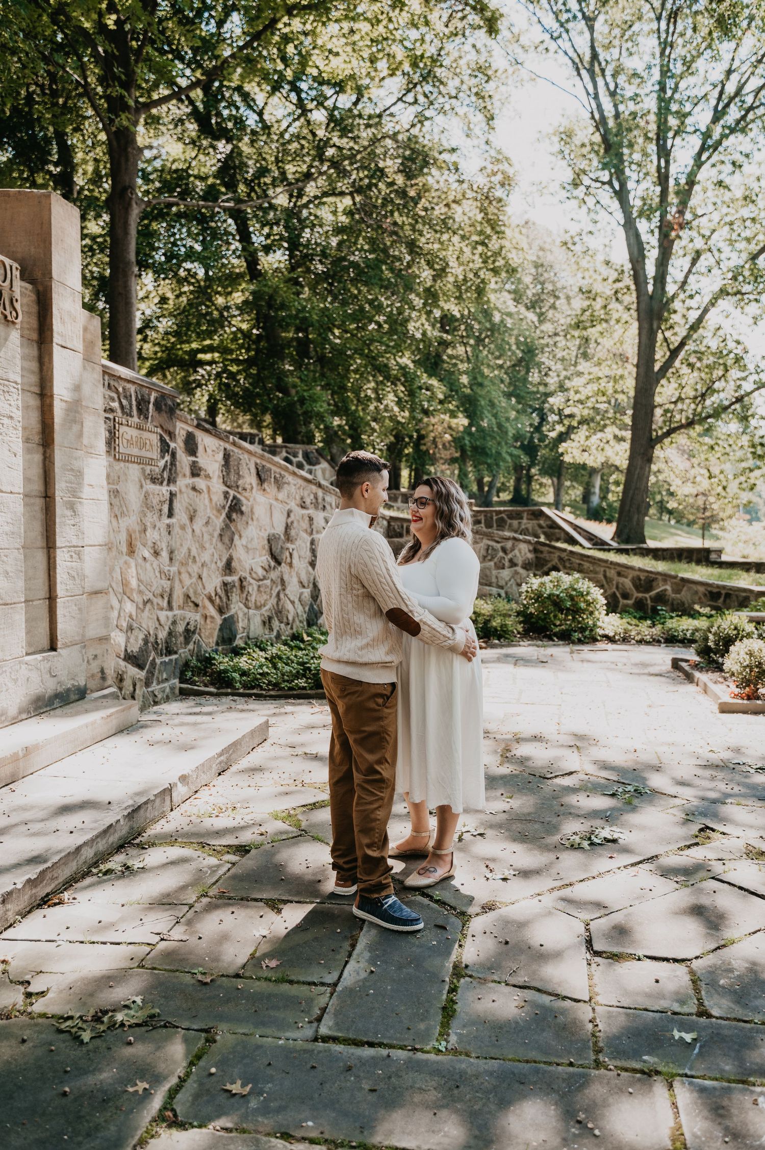 A couple embraces in a sunlit stone courtyard surrounded by lush greenery and architectural details.