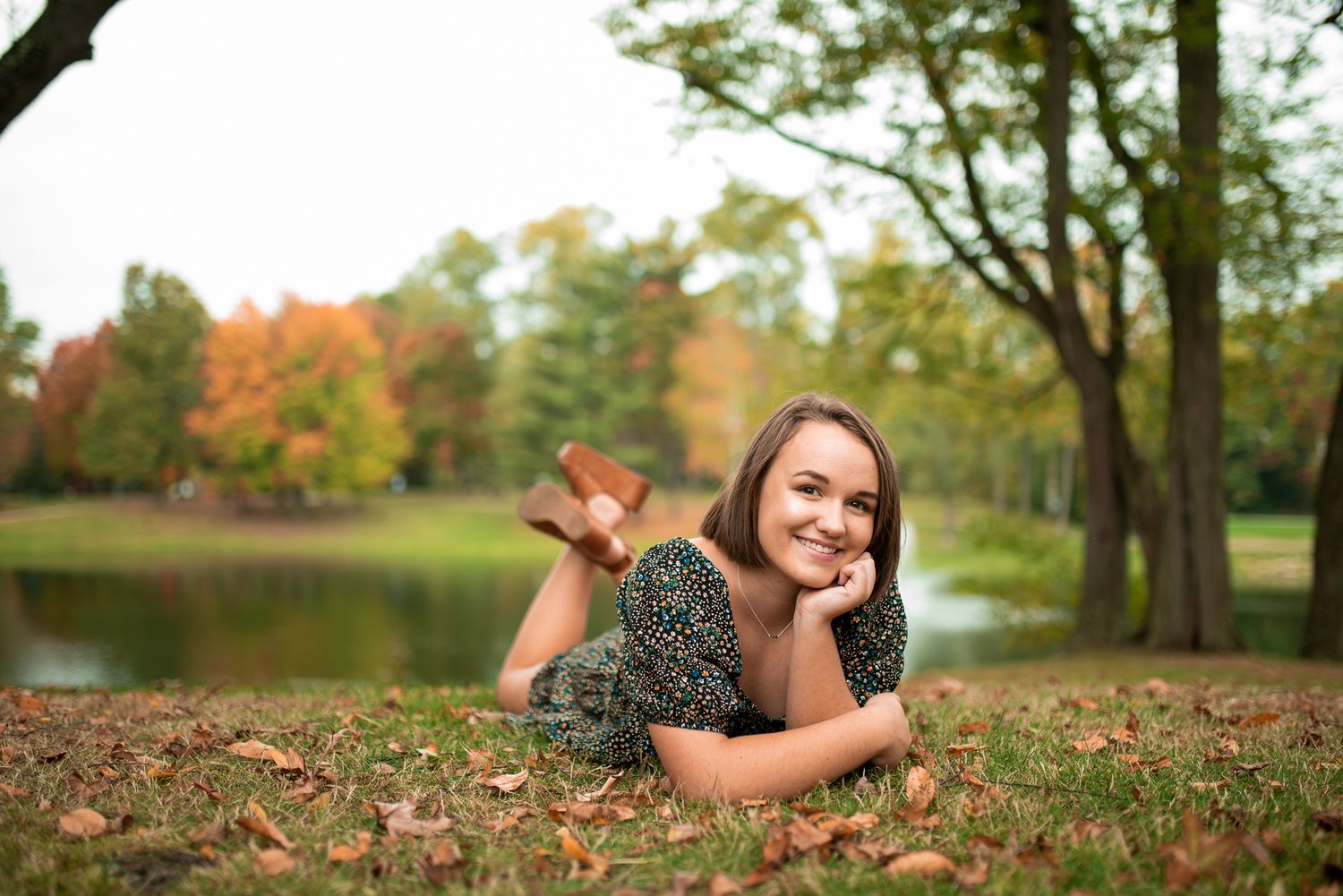 A woman lies in autumn leaves wearing casual clothing and a warm smile as fall colors surround her in a park setting.