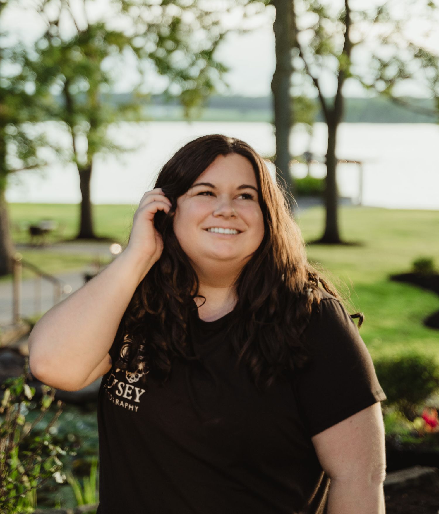Smiling young woman with dark hair wearing a black t-shirt, standing outdoors near a lake surrounded by green trees.