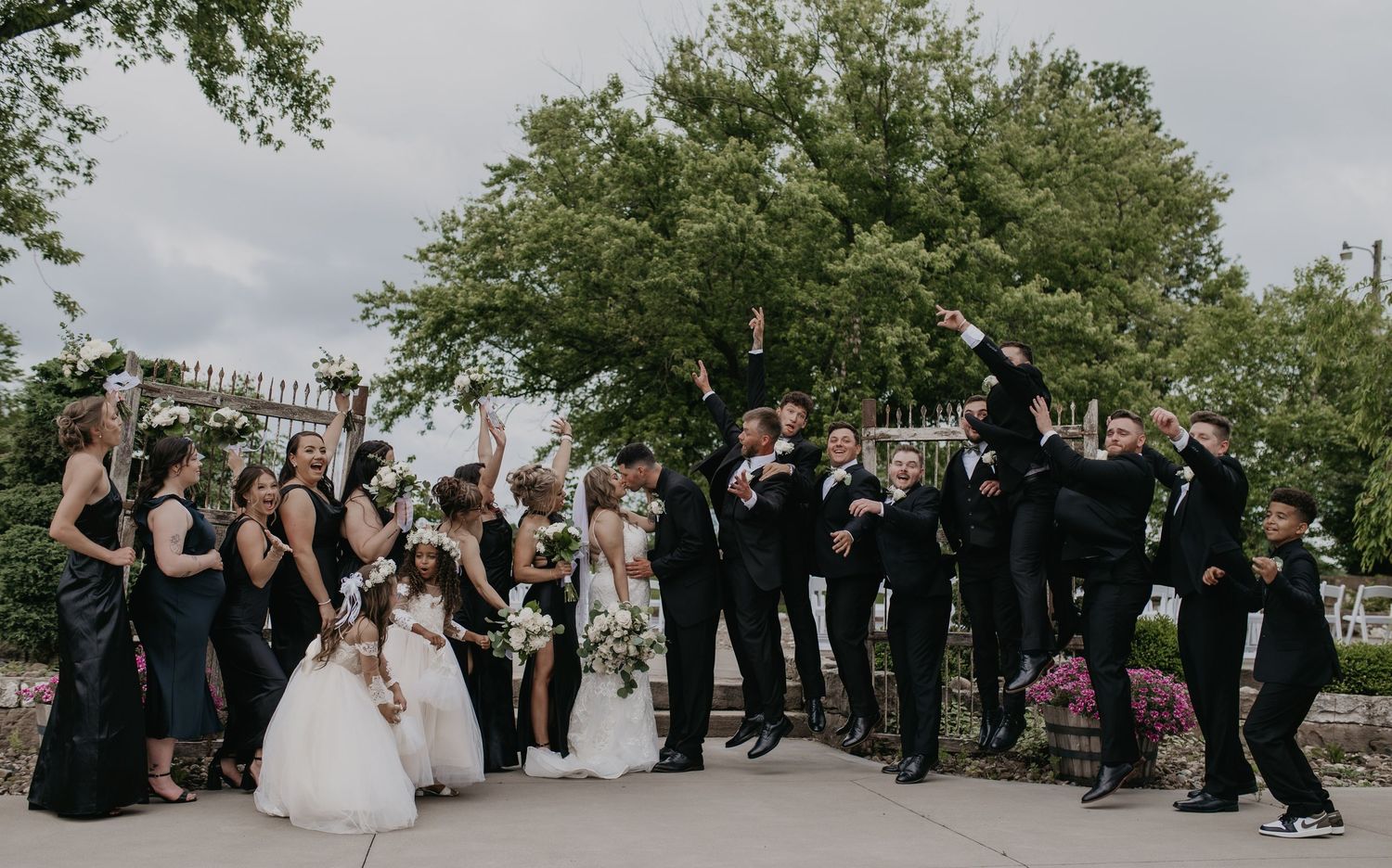 Wedding party and guests gather outdoors under green trees for a group photo celebration.