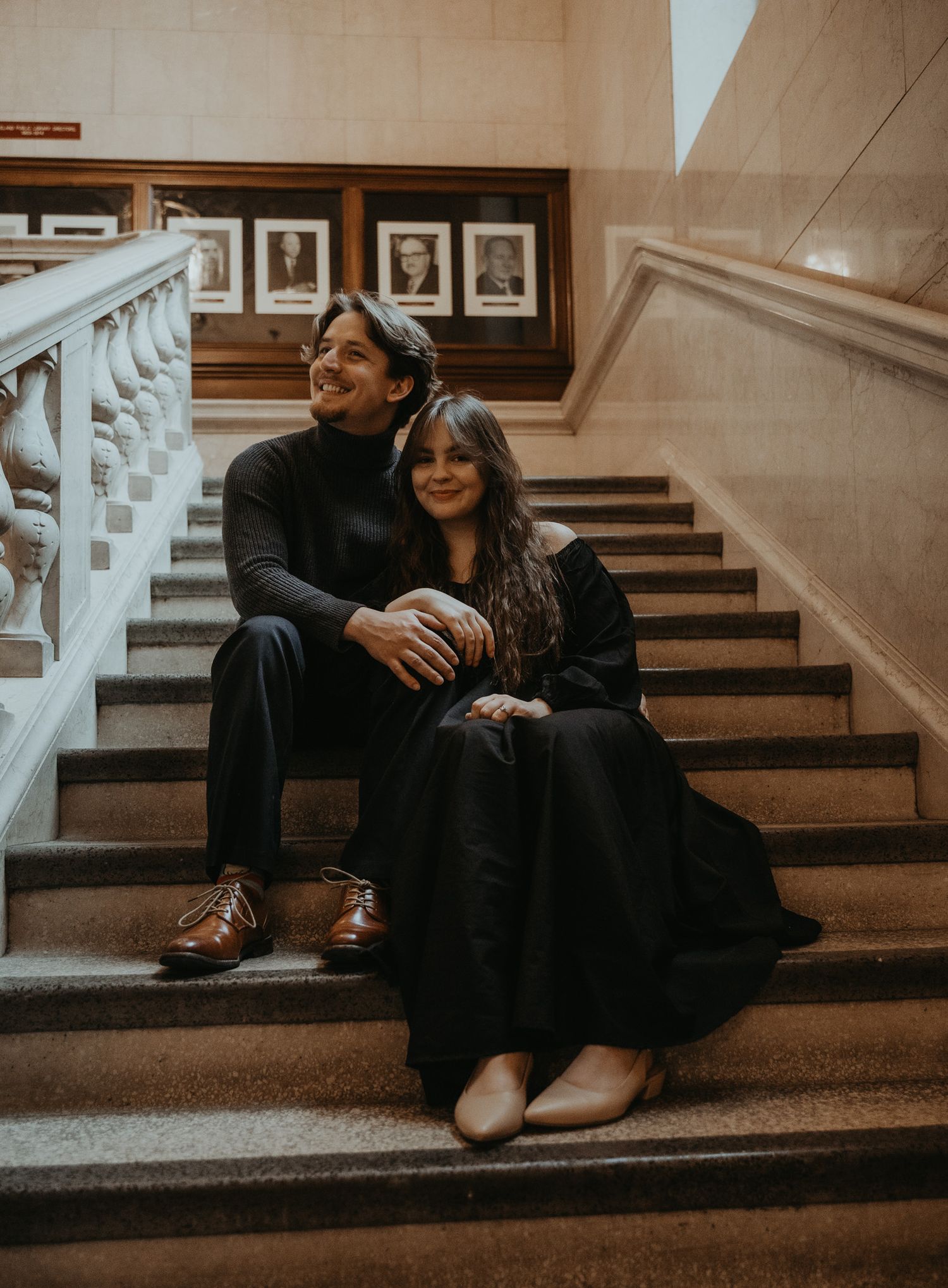 A couple sits intimately together on carpeted stairs in a dimly lit hallway with framed photos on the wall.