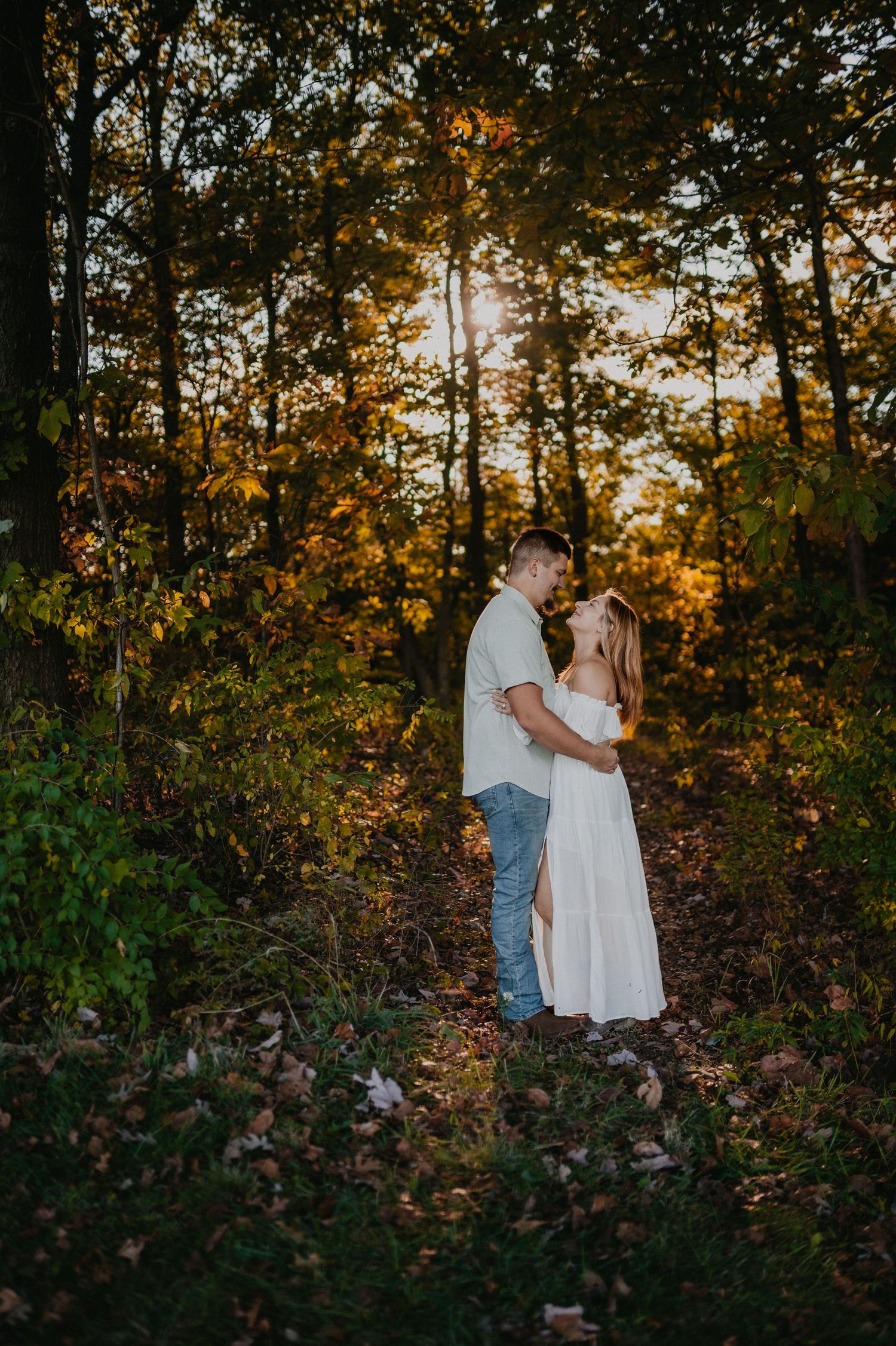 A couple in white and casual attire shares a romantic moment against an autumn forest backdrop at sunset.