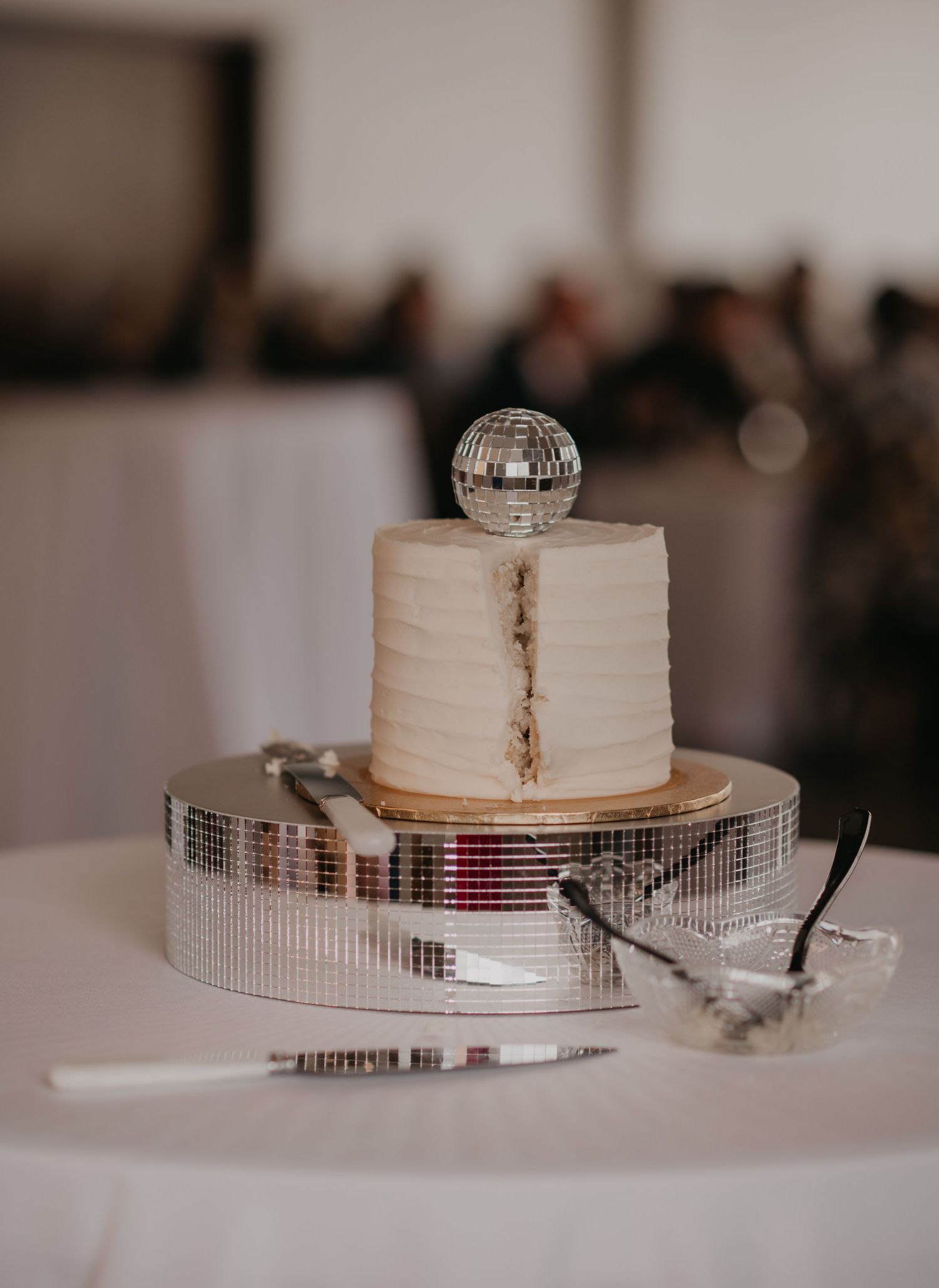 White wedding cake with simple textured frosting displayed on a mirrored stand with serving utensils.