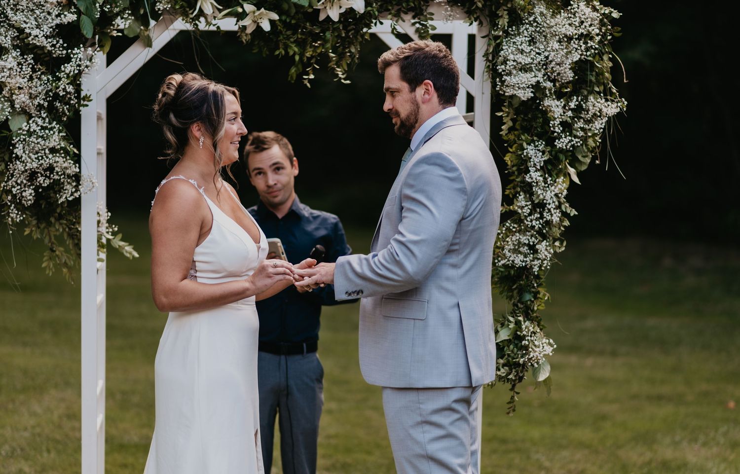 A couple exchanges rings during an outdoor wedding ceremony surrounded by white floral arrangements and greenery.