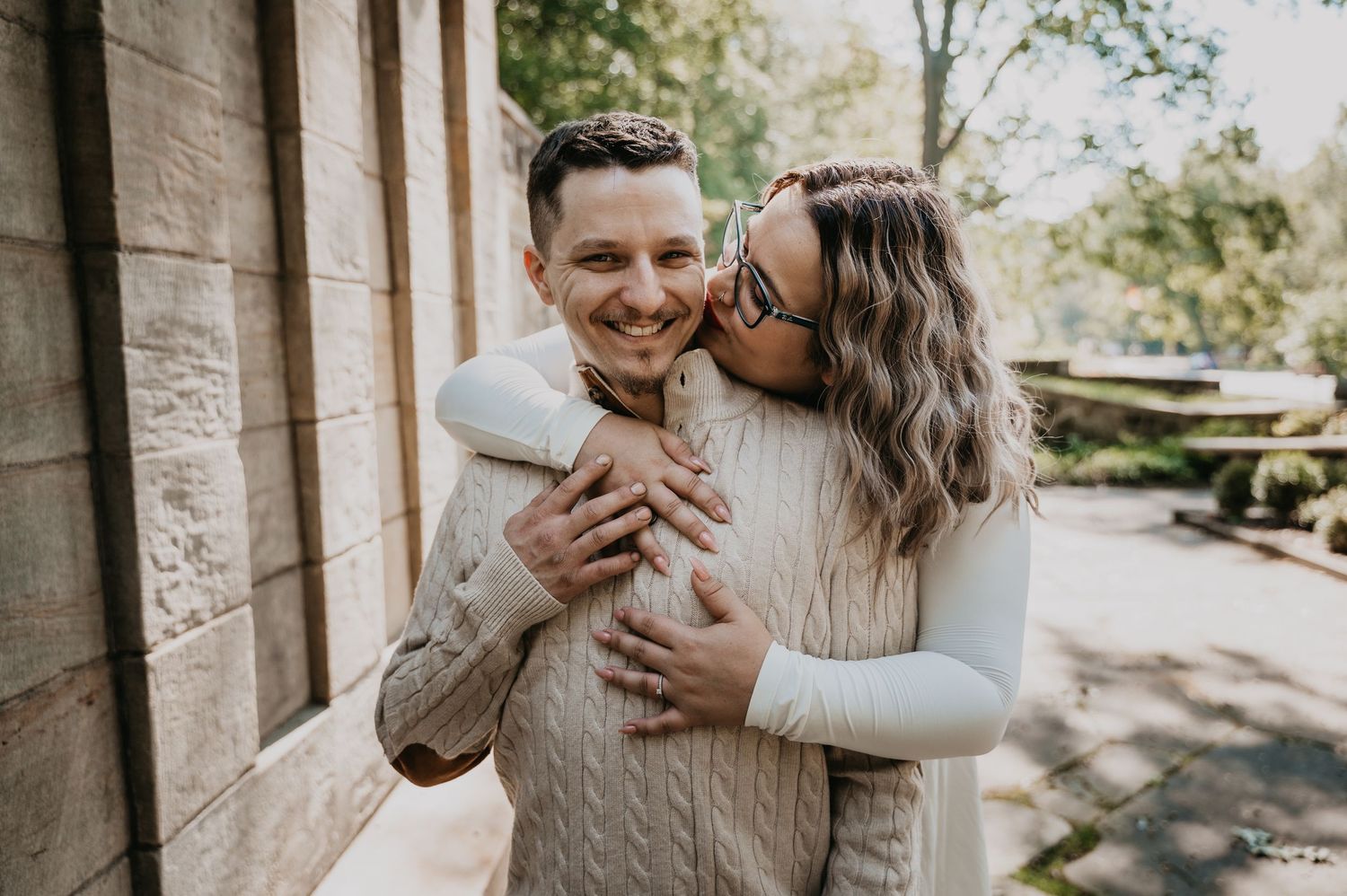 Couple embracing and laughing together outdoors on a sunny day near stone architecture.