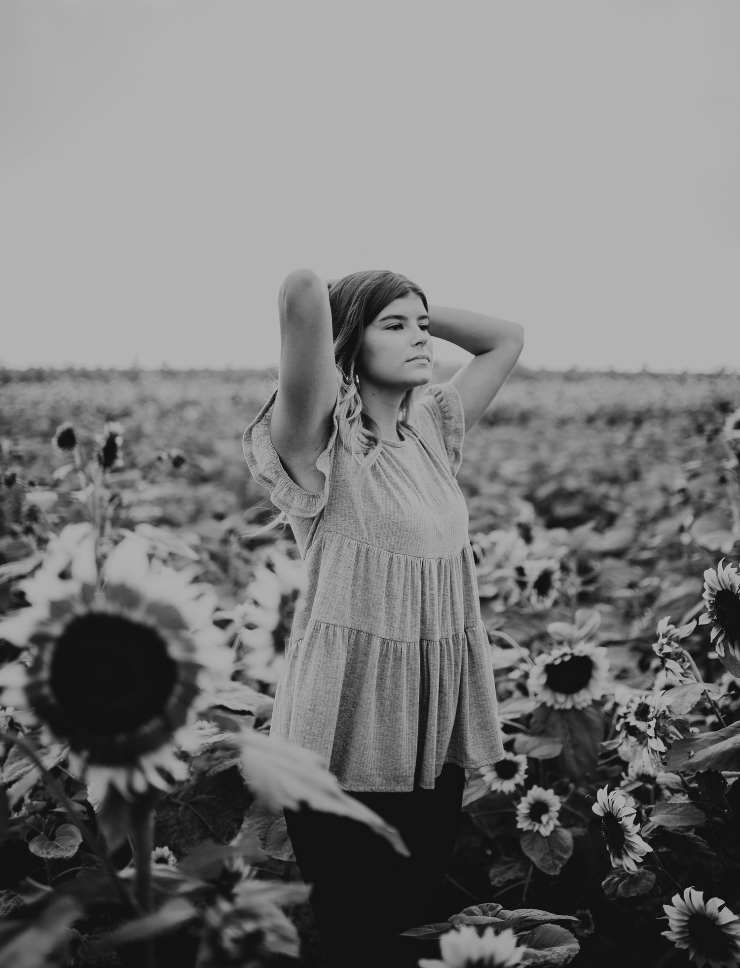 Black and white artistic portrait of a figure in a flowing dress standing among blooming sunflowers in a field at sunset.