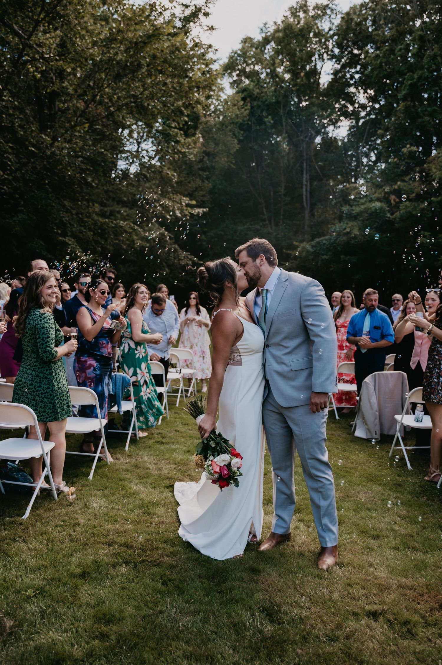 Wedding guests gather on a grassy lawn with white chairs during an outdoor reception celebration.
