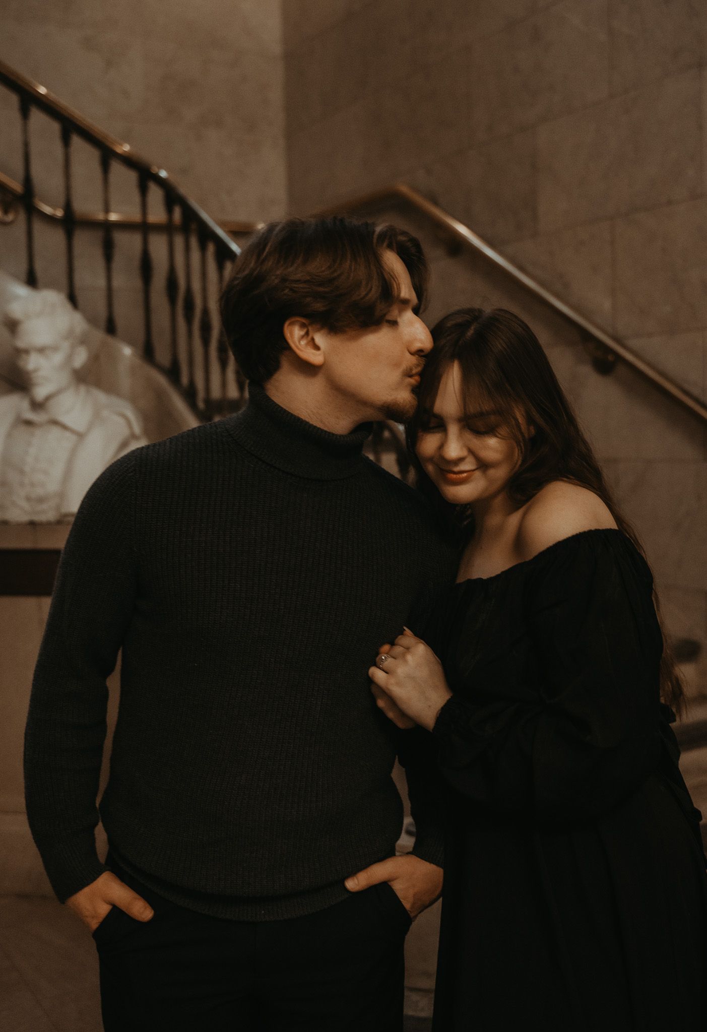 A romantic couple in matching black sweaters share an intimate moment on a staircase in moody lighting.