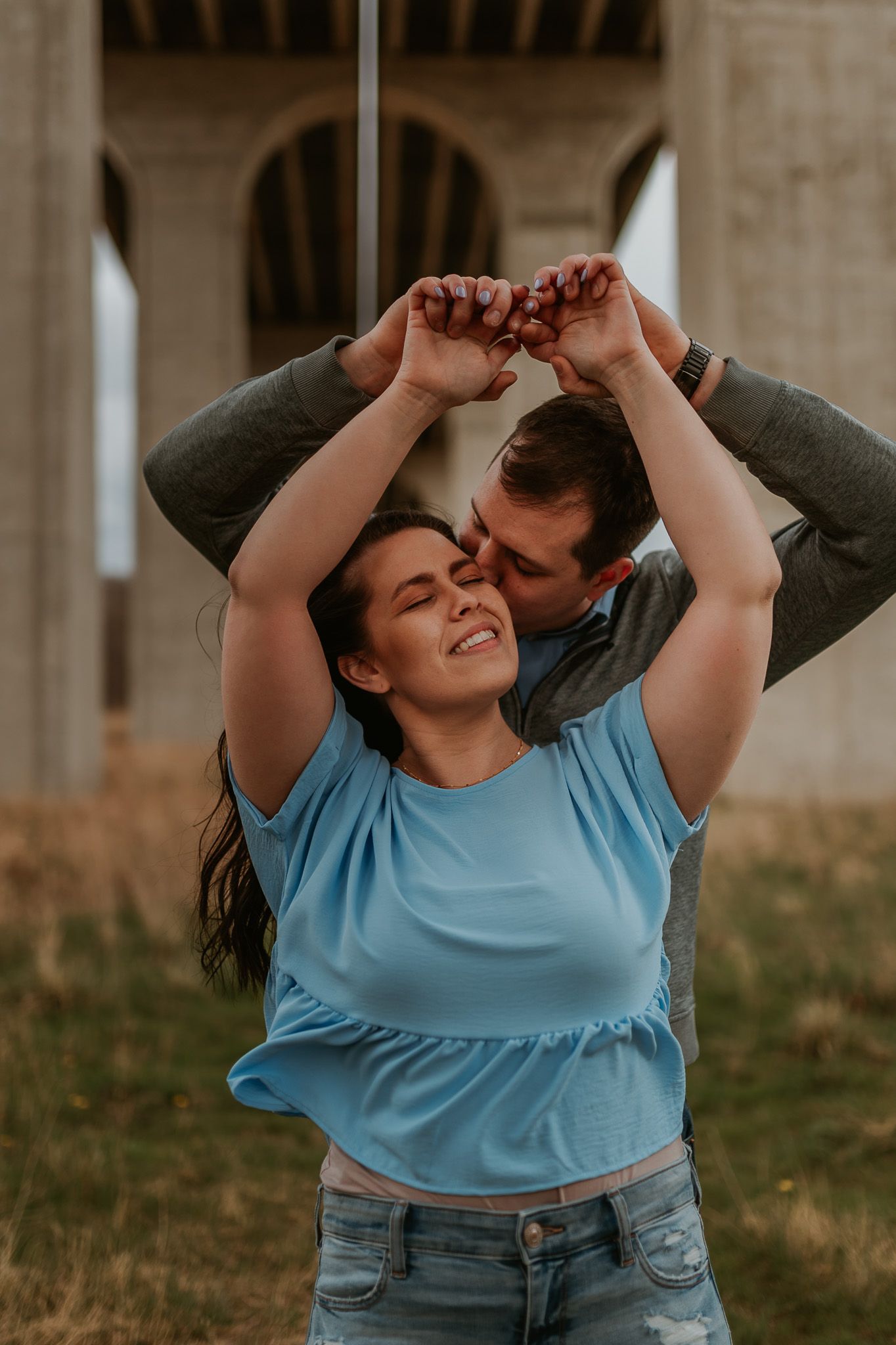 A couple shares a playful and romantic moment under a bridge structure during a photoshoot.