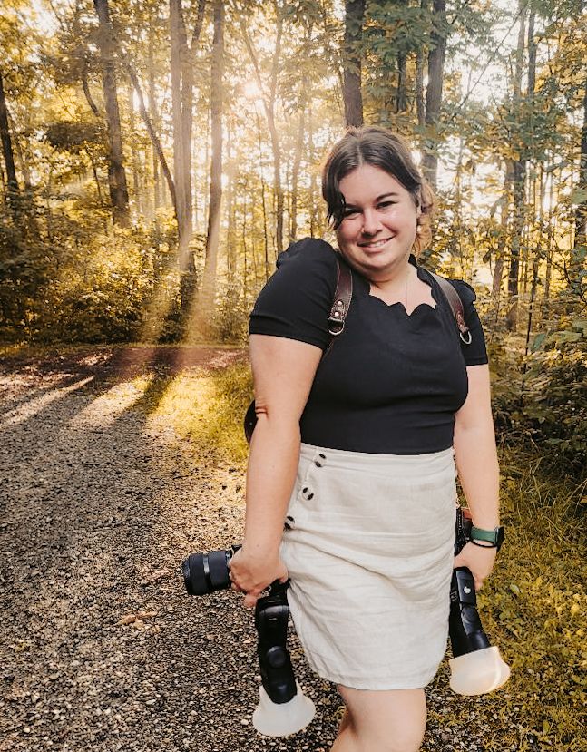 Someone standing on a wooded trail holding photography equipment in warm golden lighting.