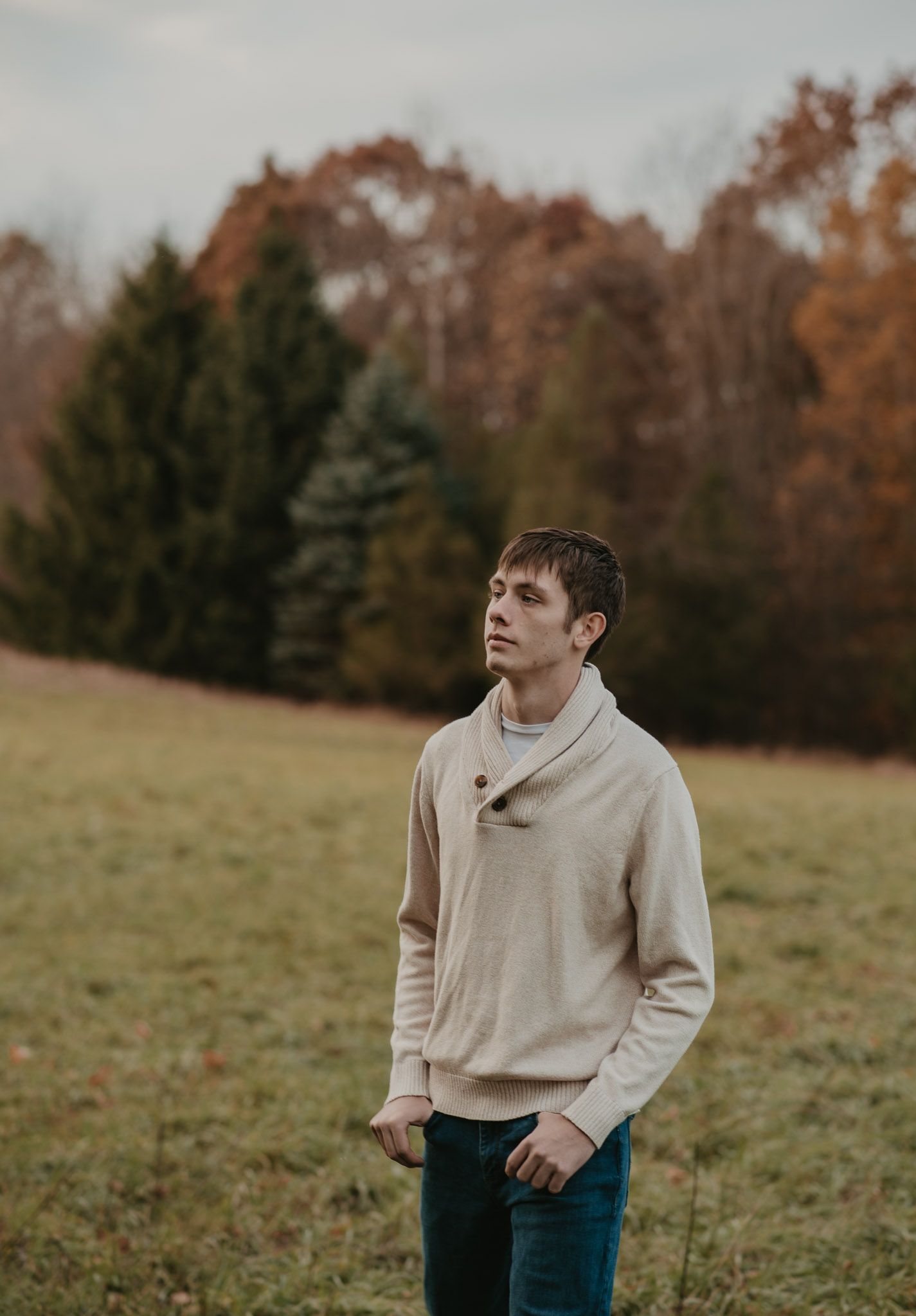 A person in a beige sweater stands in an autumn field with trees in the background.