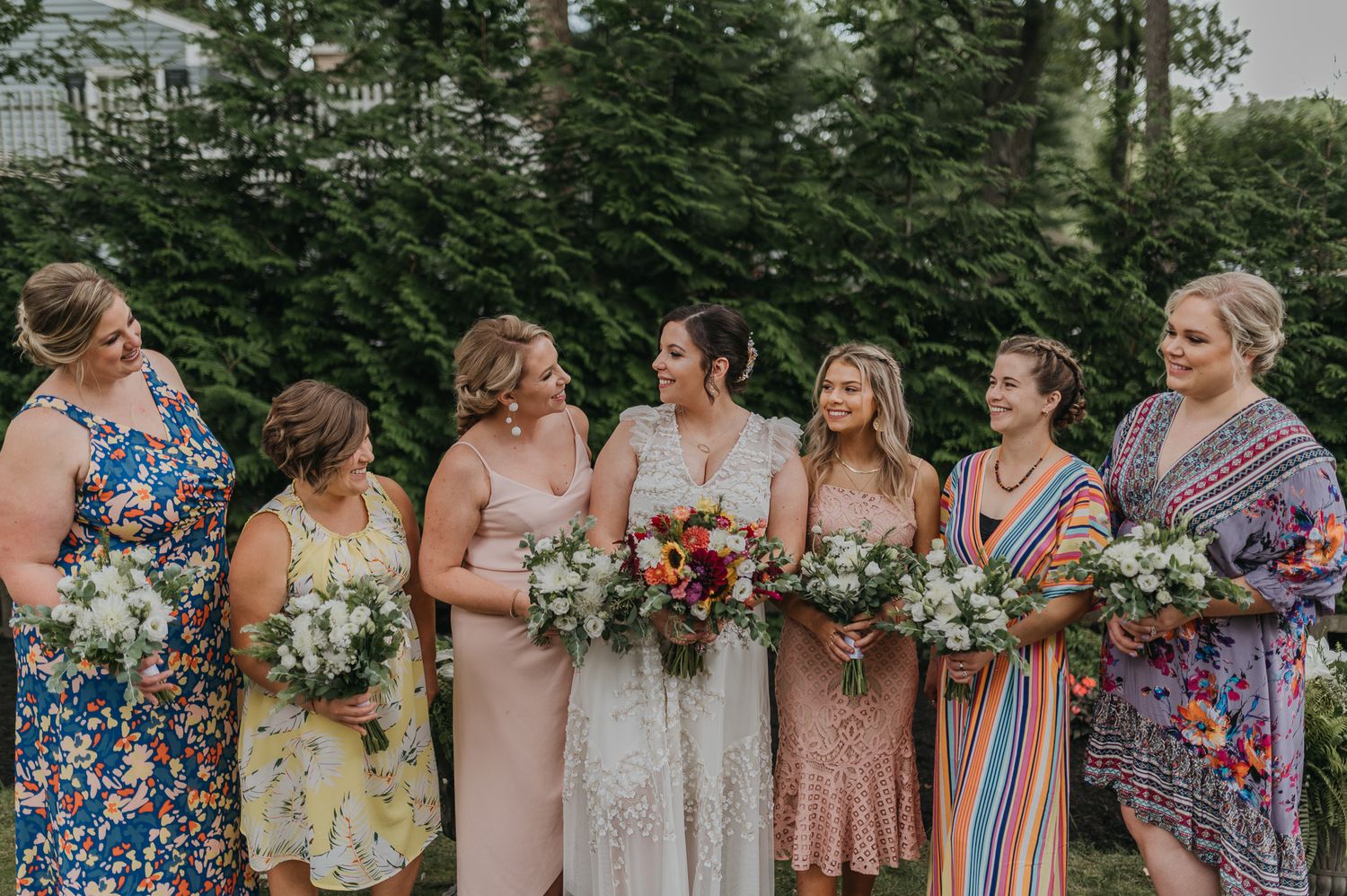 Row of bridesmaids holding colorful floral bouquets outdoors at summer wedding.