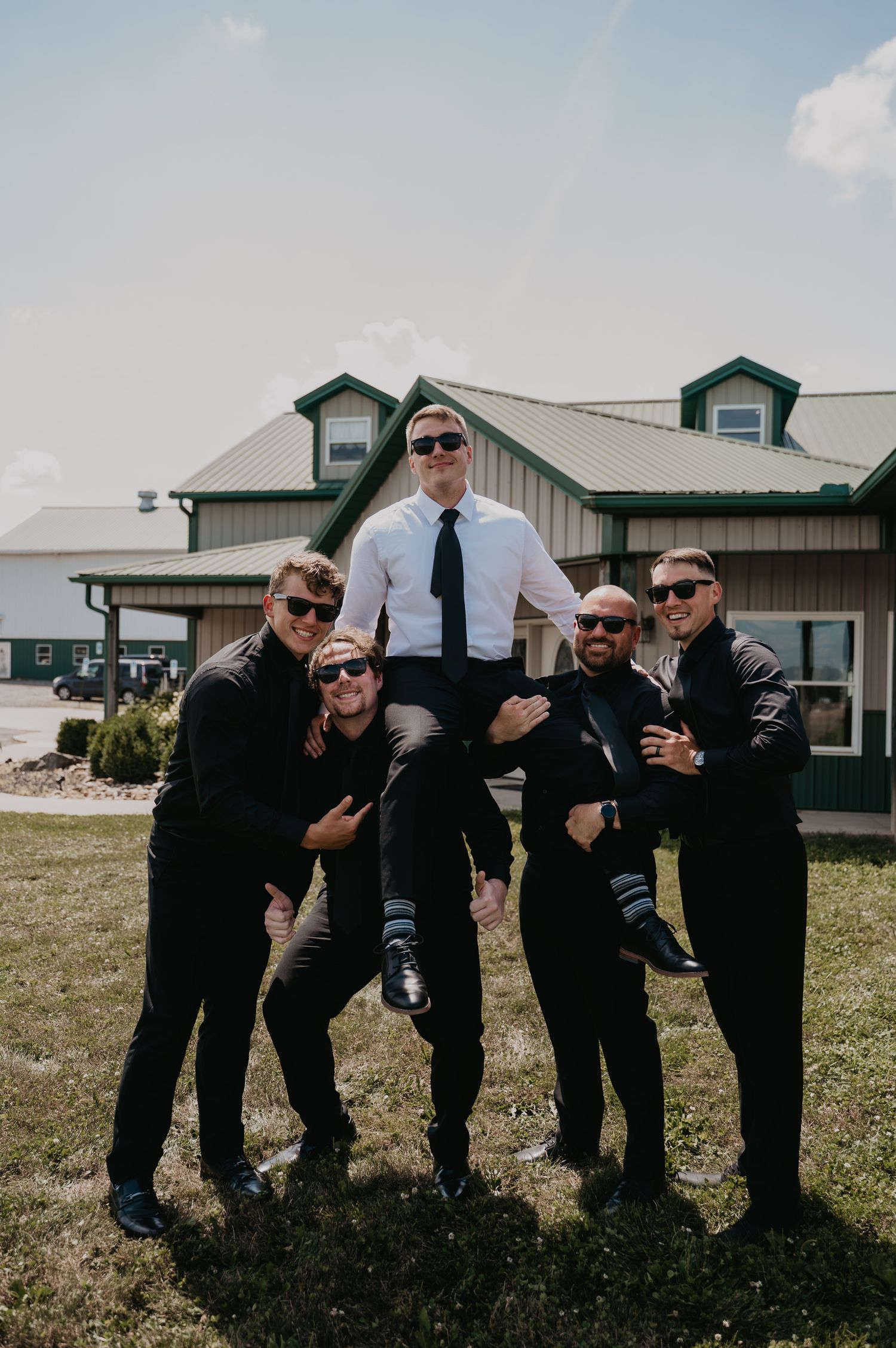 Groomsmen in black suits and sunglasses pose playfully in a line on grass with buildings in background.
