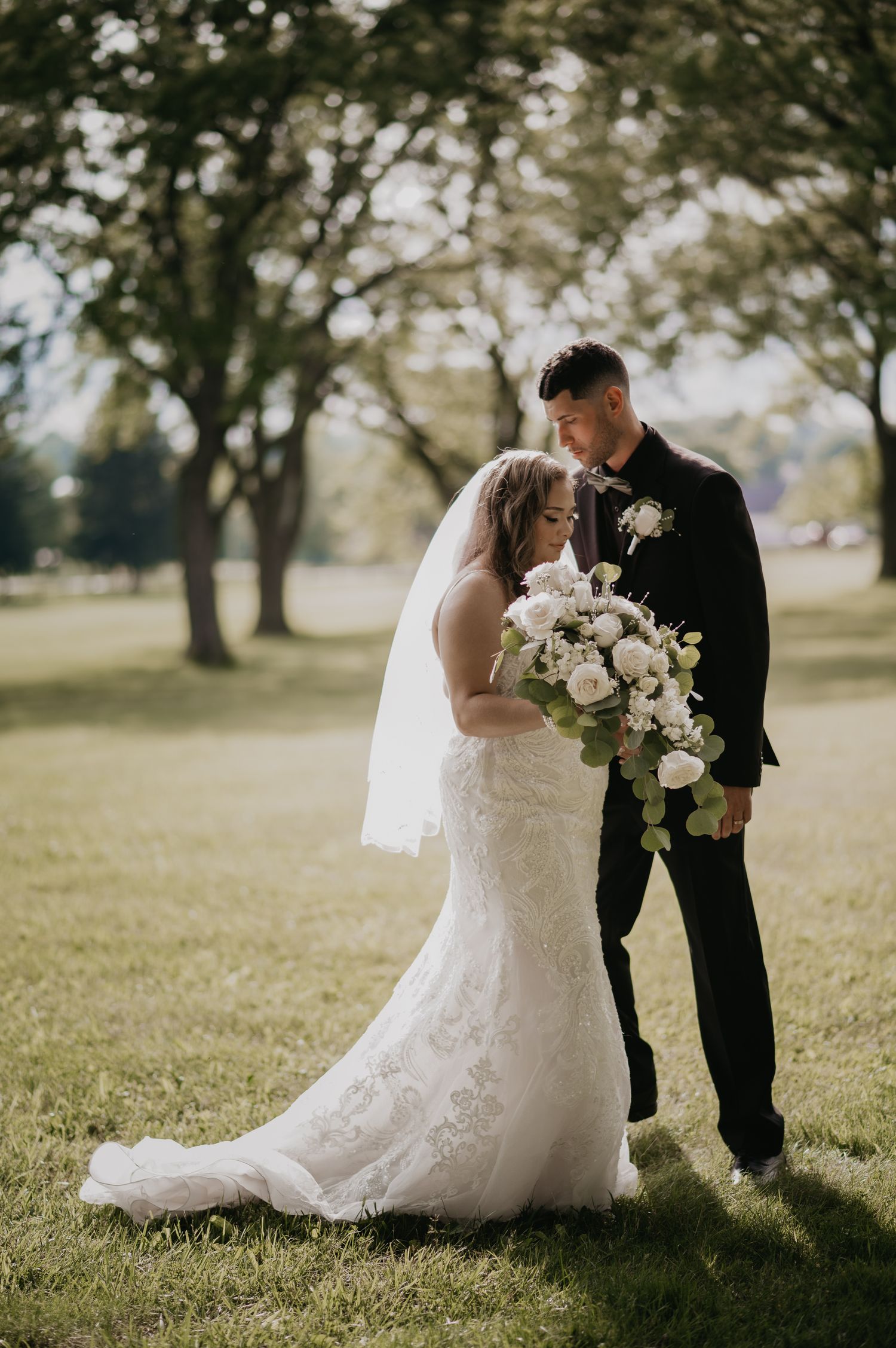 Romantic wedding couple poses in silhouette against sunlit grassy field with trees.