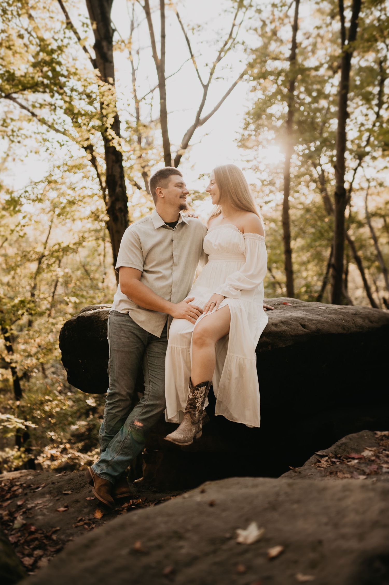 A couple sits together on a large rock formation surrounded by autumn foliage.