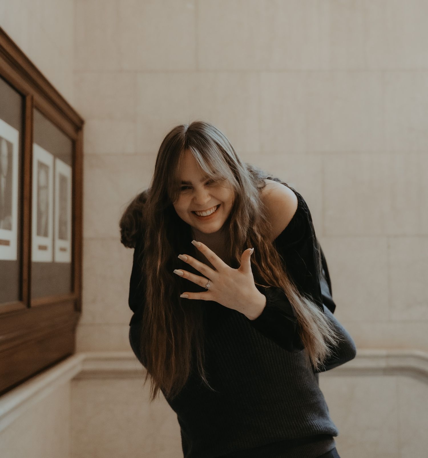 Person in a black sweater smiles and moves playfully against a beige wall with framed artwork.