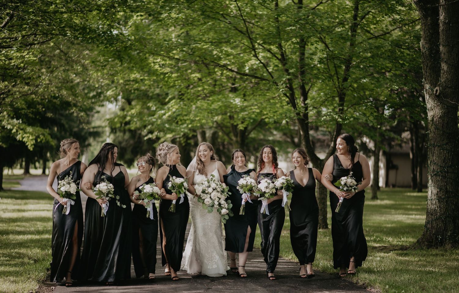 Wedding party in long black dresses and white gown pose together under green trees in outdoor setting.