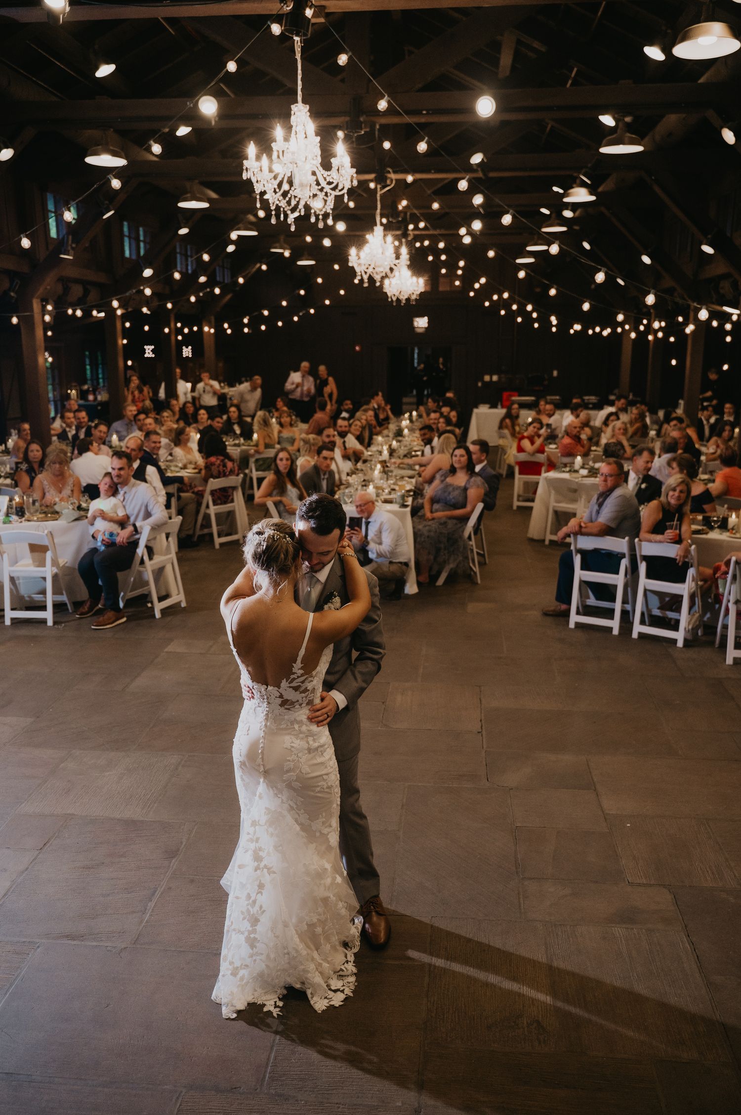 Couple shares their first dance at an elegant indoor wedding reception with string lights and chandeliers overhead.