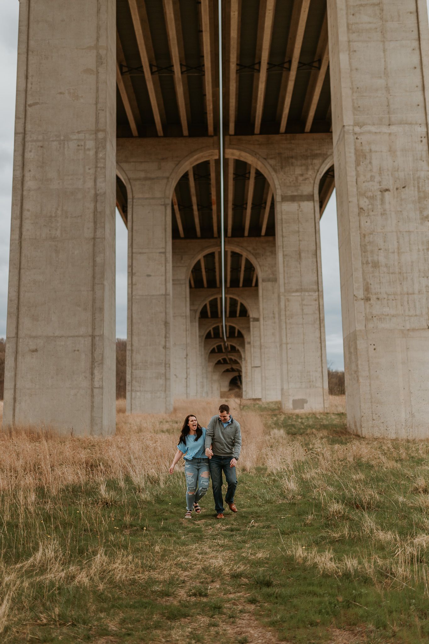 A couple walks hand in hand beneath massive concrete bridge pillars on a grassy hill.