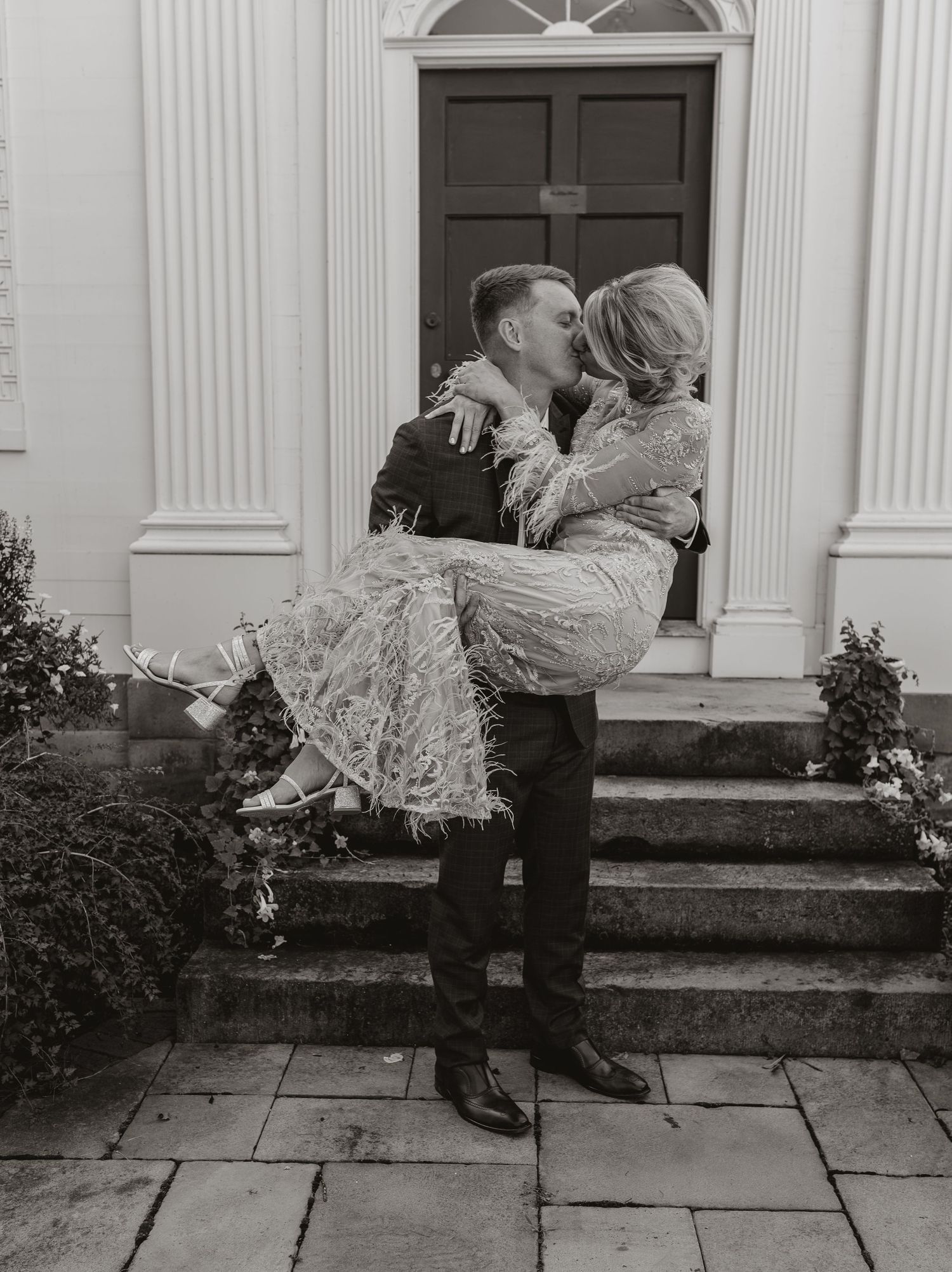 Black and white photo of a groom carrying his bride in front of an elegant doorway.