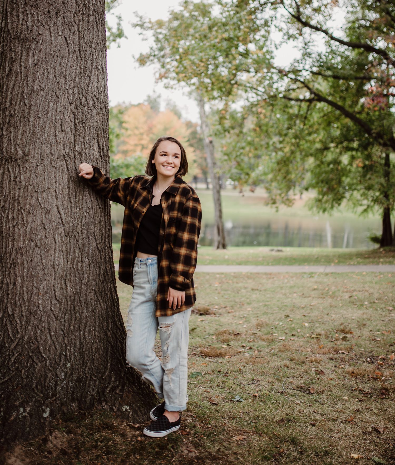Casual autumn outdoor portrait with plaid flannel shirt and light wash jeans standing beside large tree trunk in park setting.