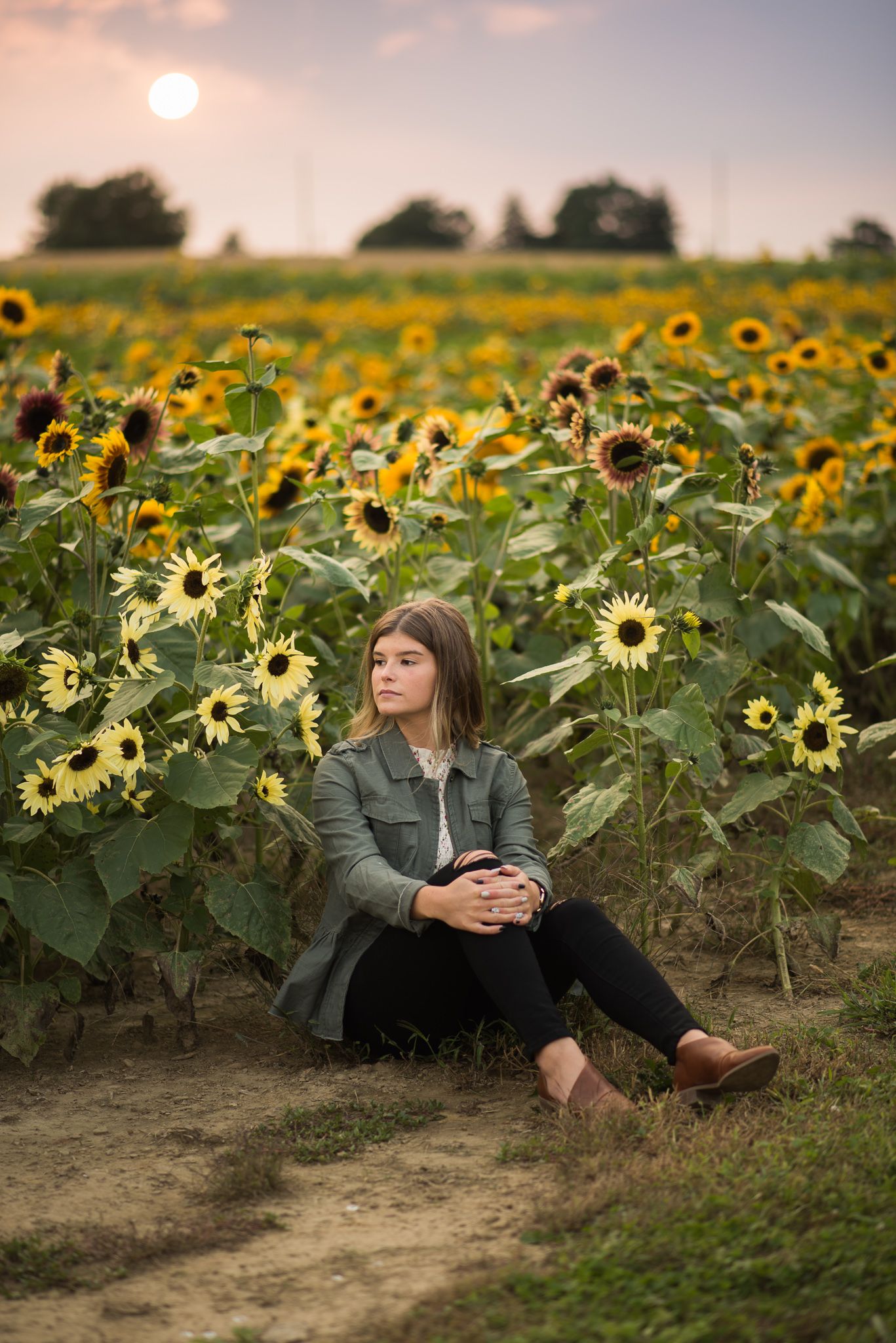 Person in green jacket sitting in field of blooming sunflowers during sunset.