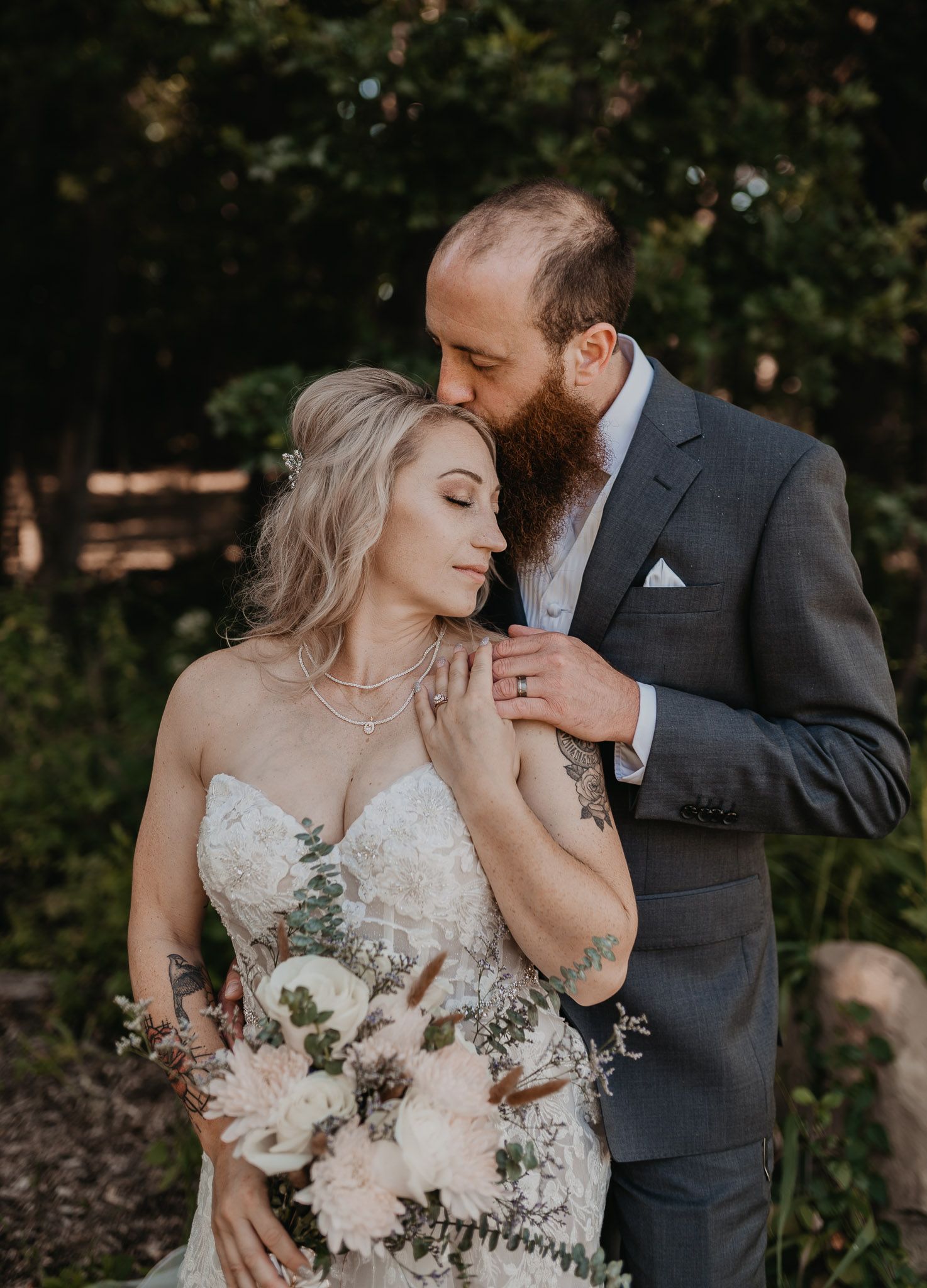 Bride in lace dress holds bouquet while groom in grey suit embraces her in outdoor garden setting.