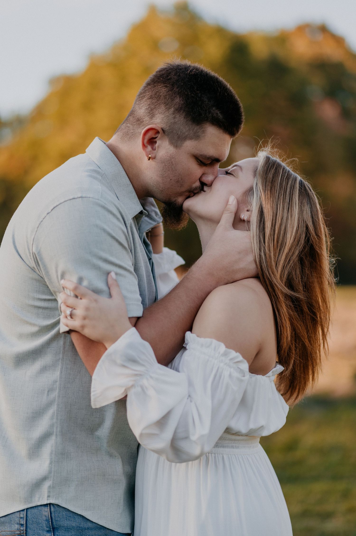 A romantic couple sharing a tender kiss outdoors during golden hour with autumn foliage in the background.