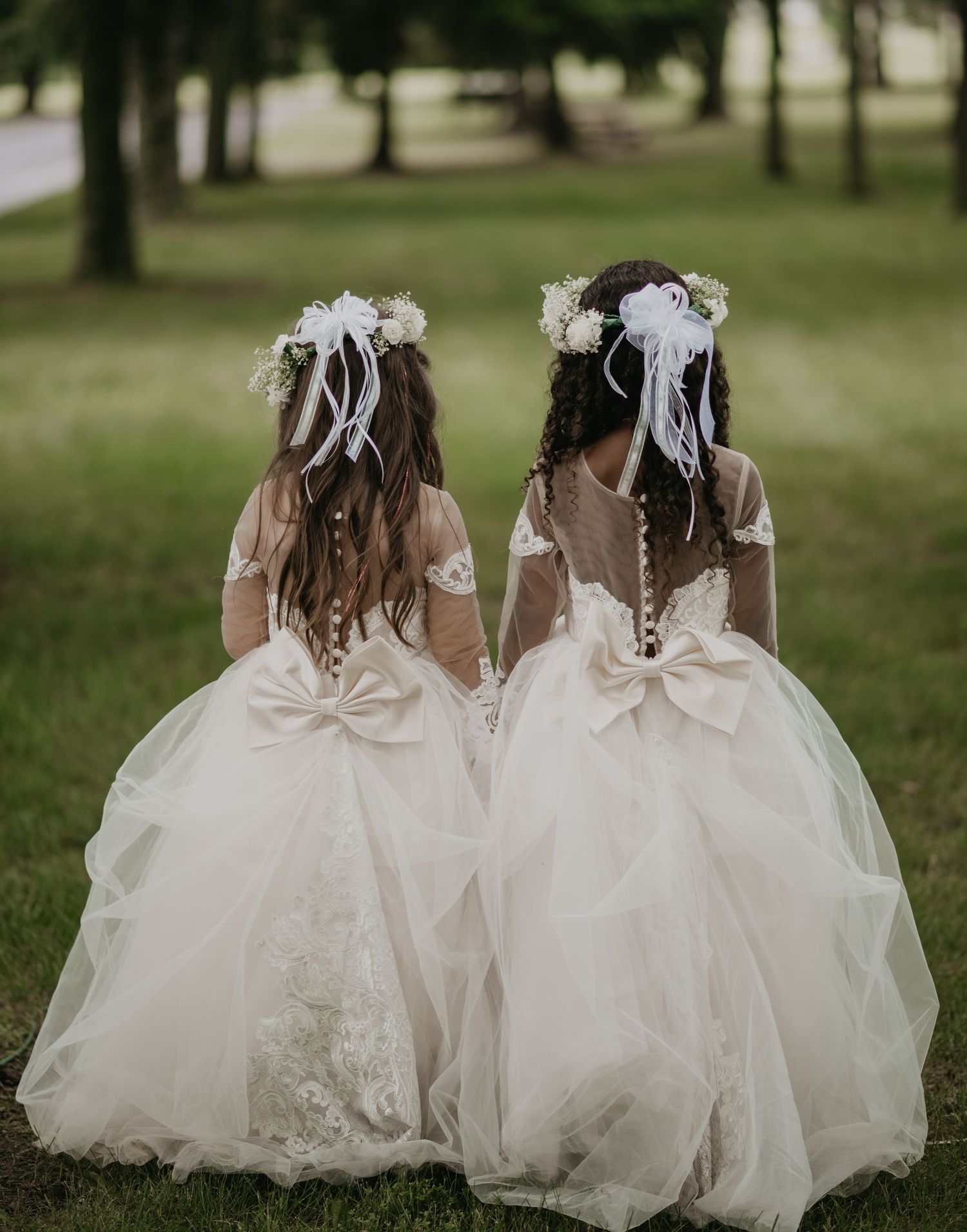 Two flower girls in matching white dresses sit together on grassy lawn during outdoor wedding.