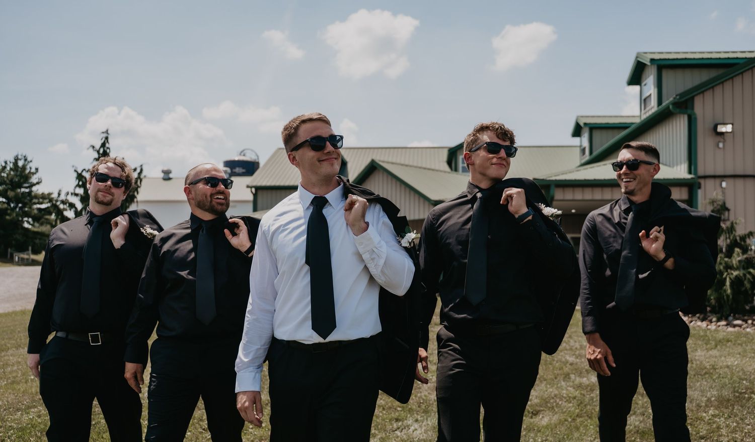 A group in formal black attire poses playfully outdoors against a rural backdrop on a sunny day.