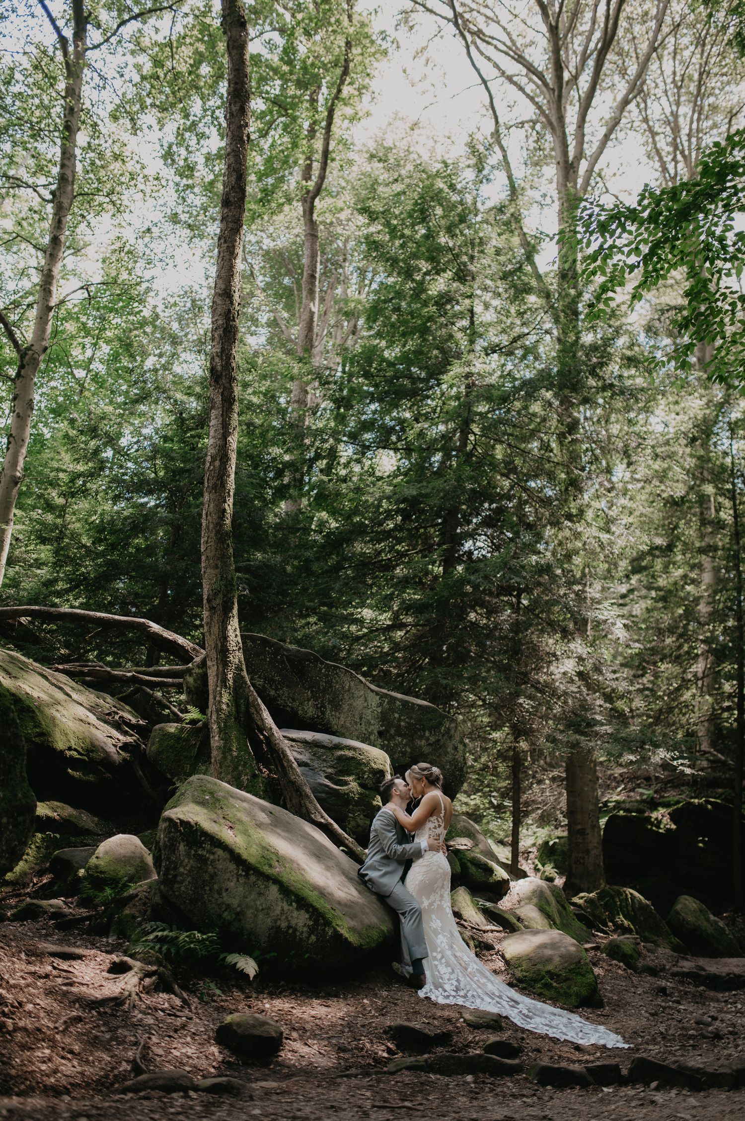 A romantic forest scene with a couple in wedding attire surrounded by moss-covered rocks and tall trees.