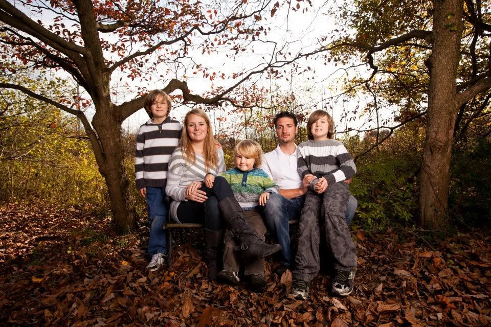 Family of five posing outdoors among autumn leaves beneath trees in a warm fall forest setting.