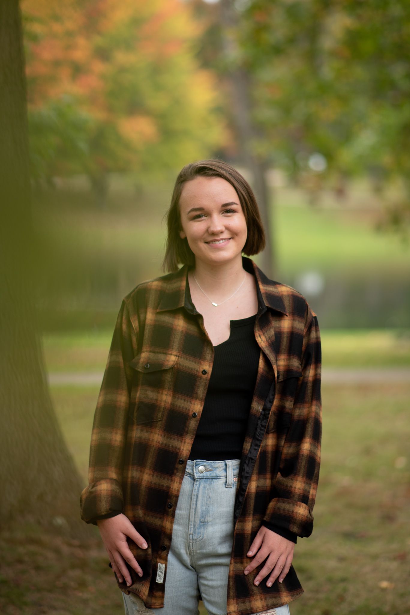 Person wearing a brown plaid flannel shirt and light jeans poses outdoors against autumn foliage in a park setting.