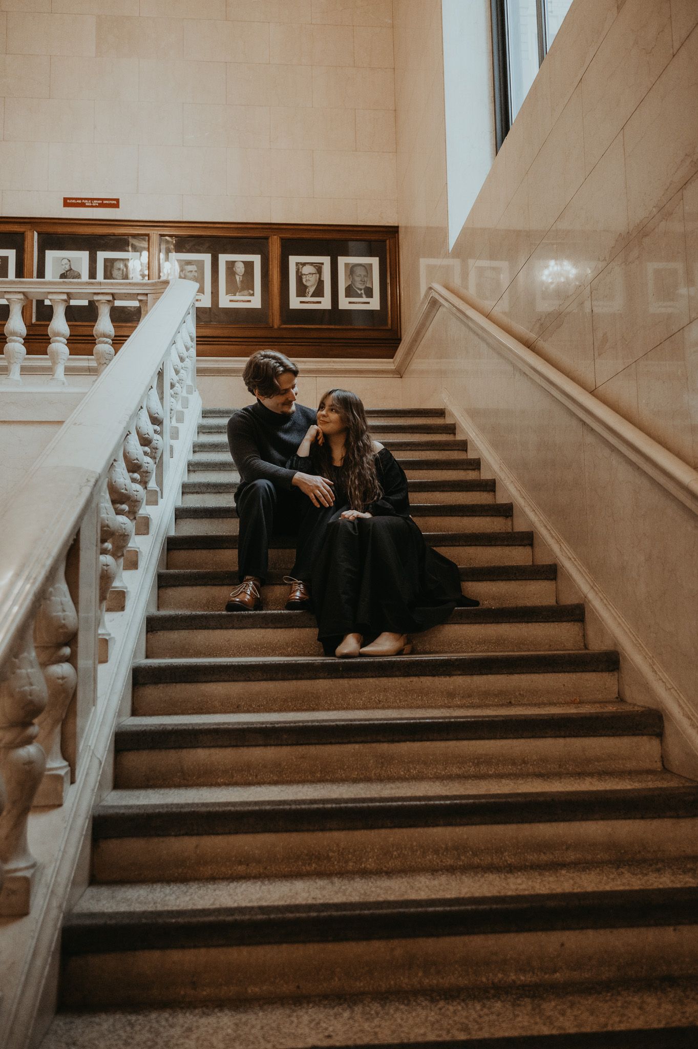 A couple sits together intimately on elegant marble stairs in a classic interior setting with framed artwork.