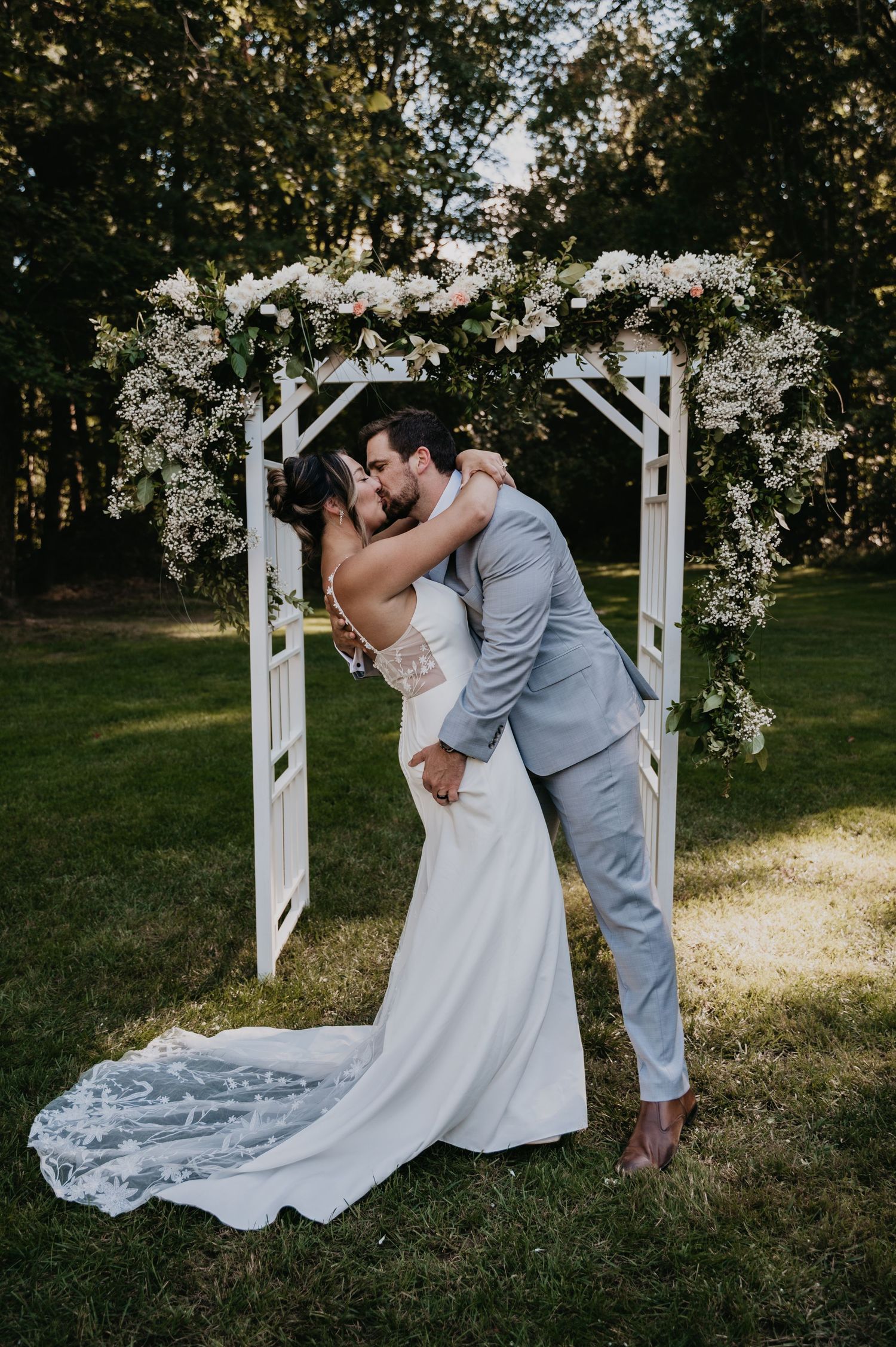 Newlyweds share a kiss under a white floral arch during their outdoor garden wedding ceremony.