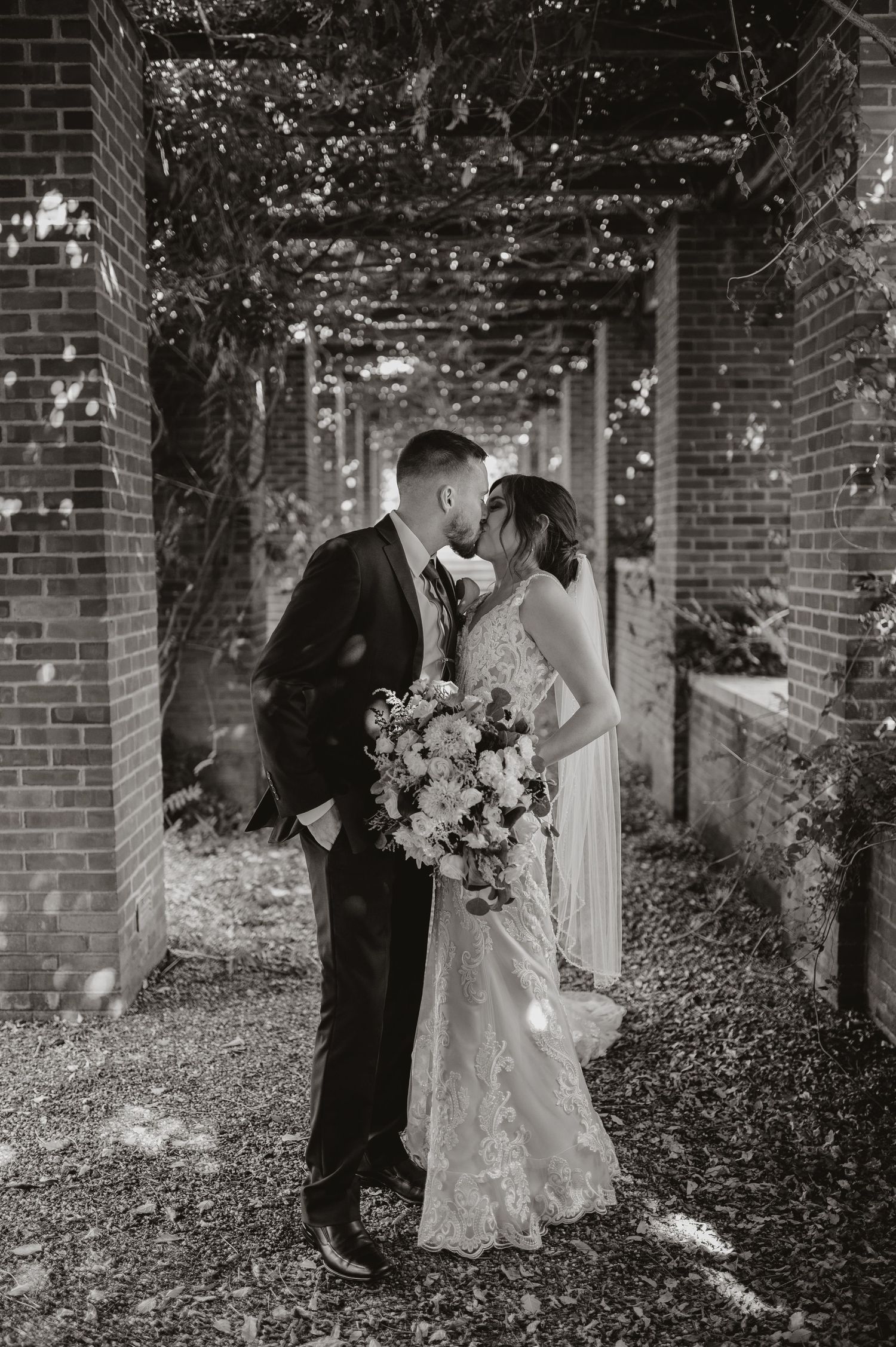 Wedding couple shares intimate moment near ivy-covered brick church entrance with vibrant floral bouquet.